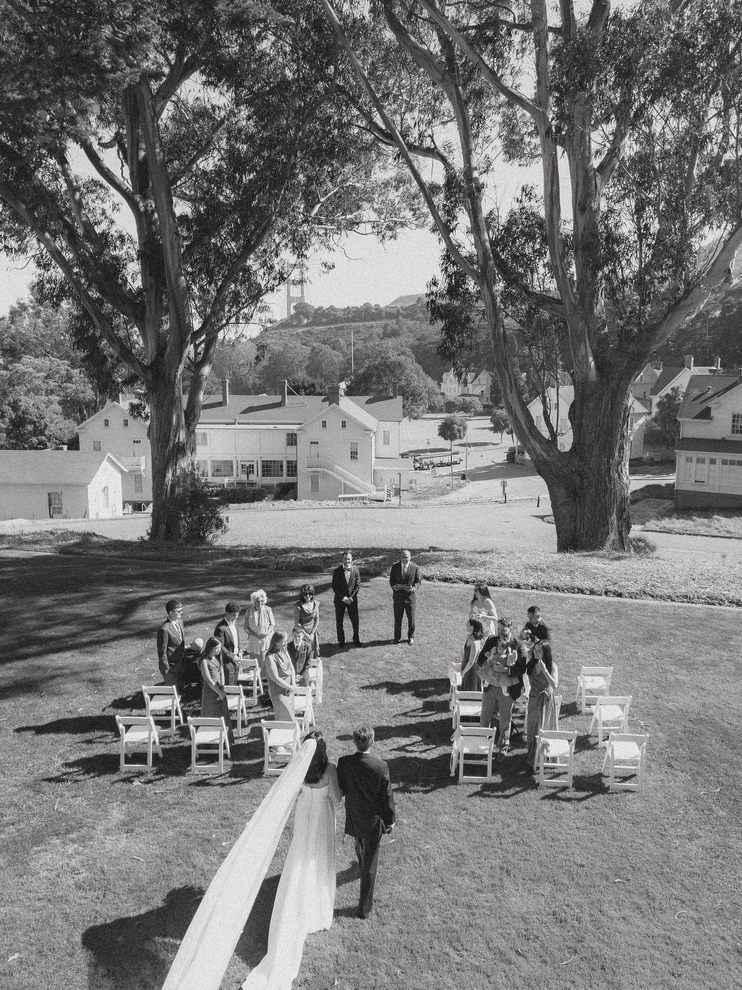 Black and white aerial view of an intimate outdoor wedding ceremony on a lawn, surrounded by large trees and buildings.