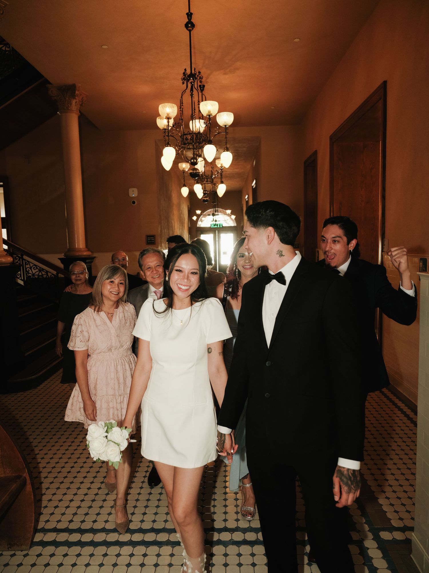 Smiling newlyweds walk through ornate hallway as guests cheer and celebrate their marriage behind them.