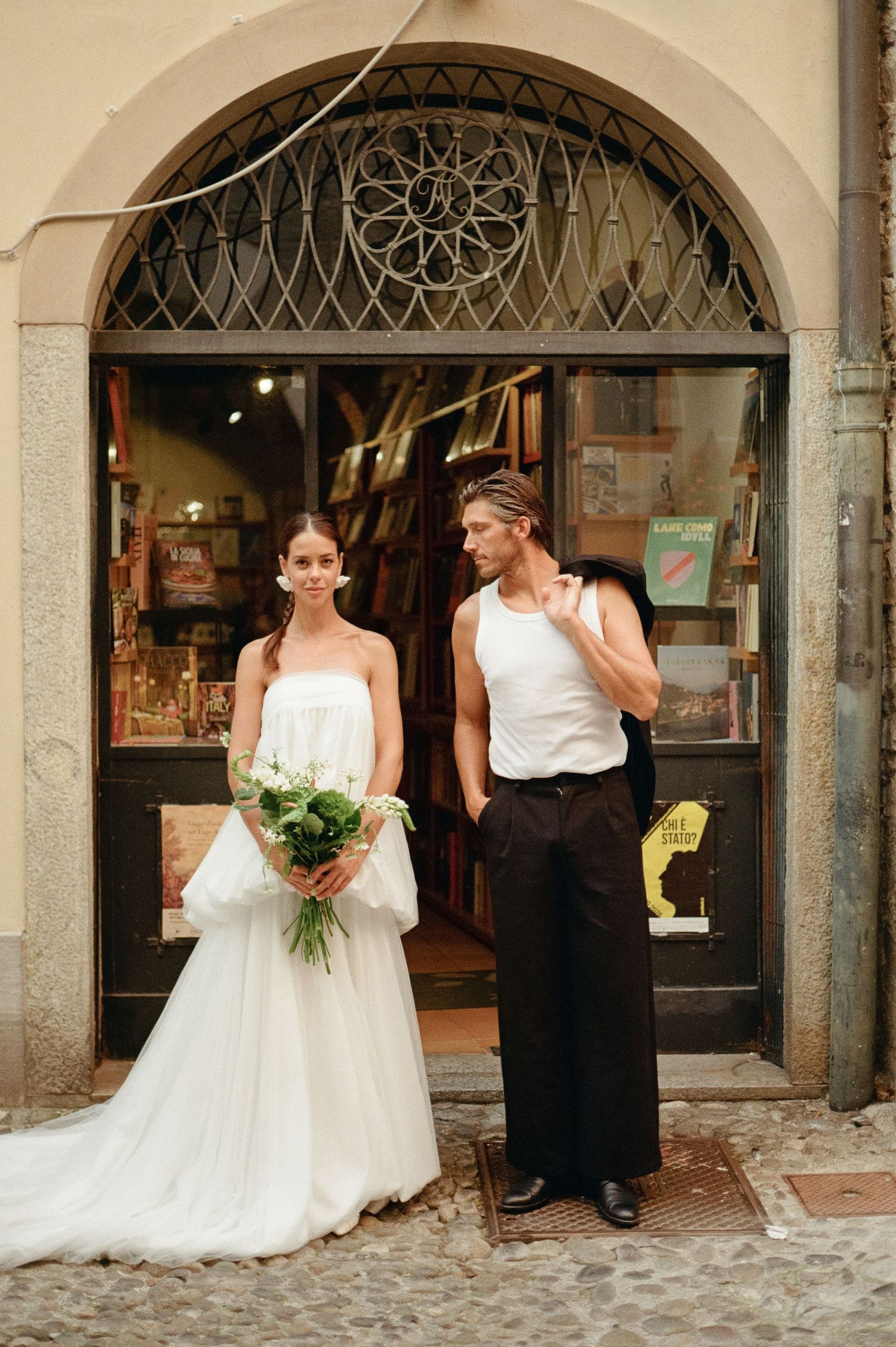 Bride holding green bouquet and groom in white tank top posing before an arched Italian bookshop.