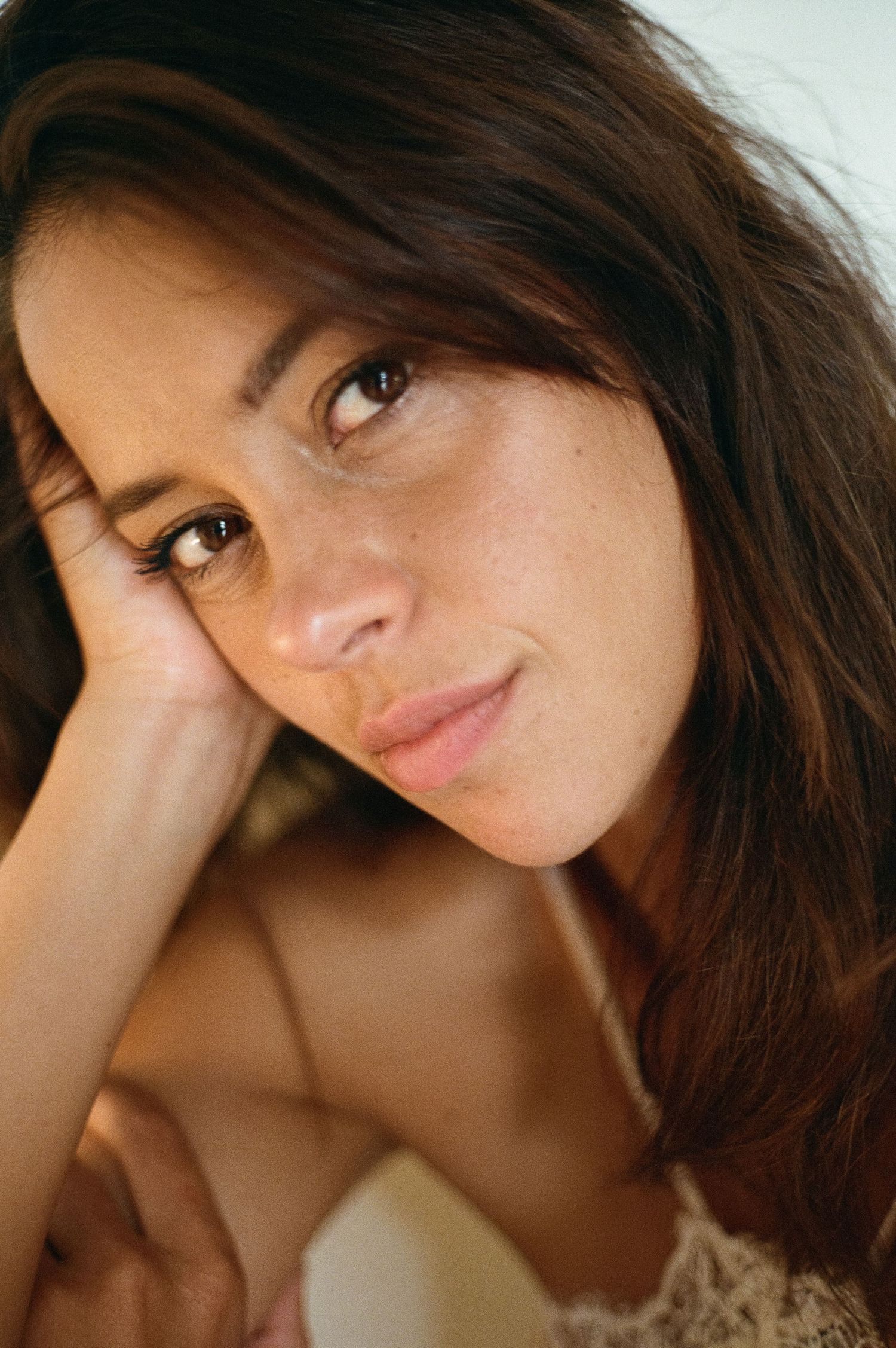 Close-up portrait of a brunette woman with freckles resting her chin on her hand, smiling softly.