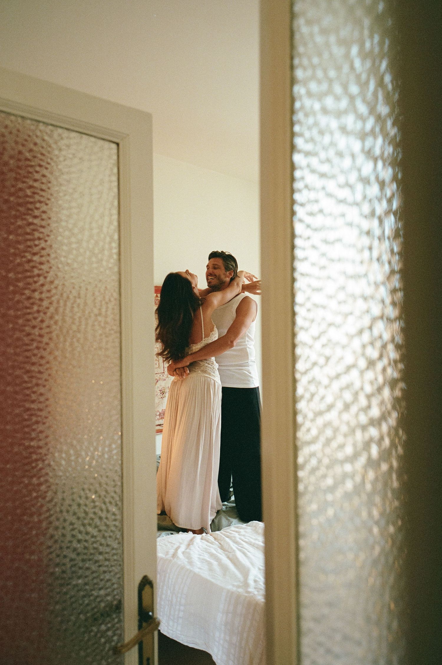 A romantic couple dances and embraces on a bed, viewed through a frosted glass door in a warmly lit bedroom.