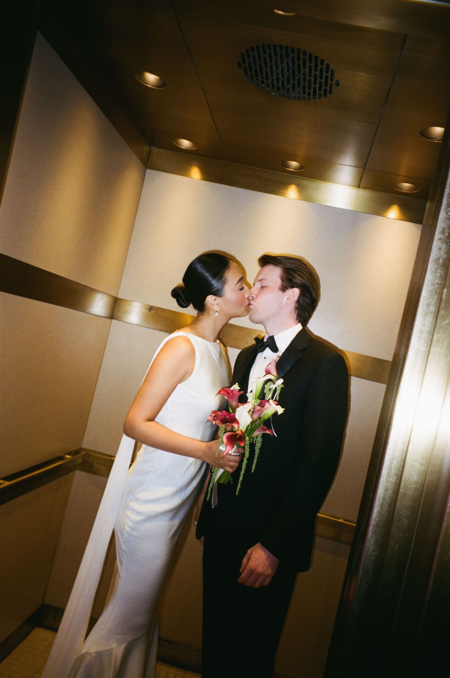 Newlyweds share a kiss inside a gold elevator, bride holding red and white floral bouquet.