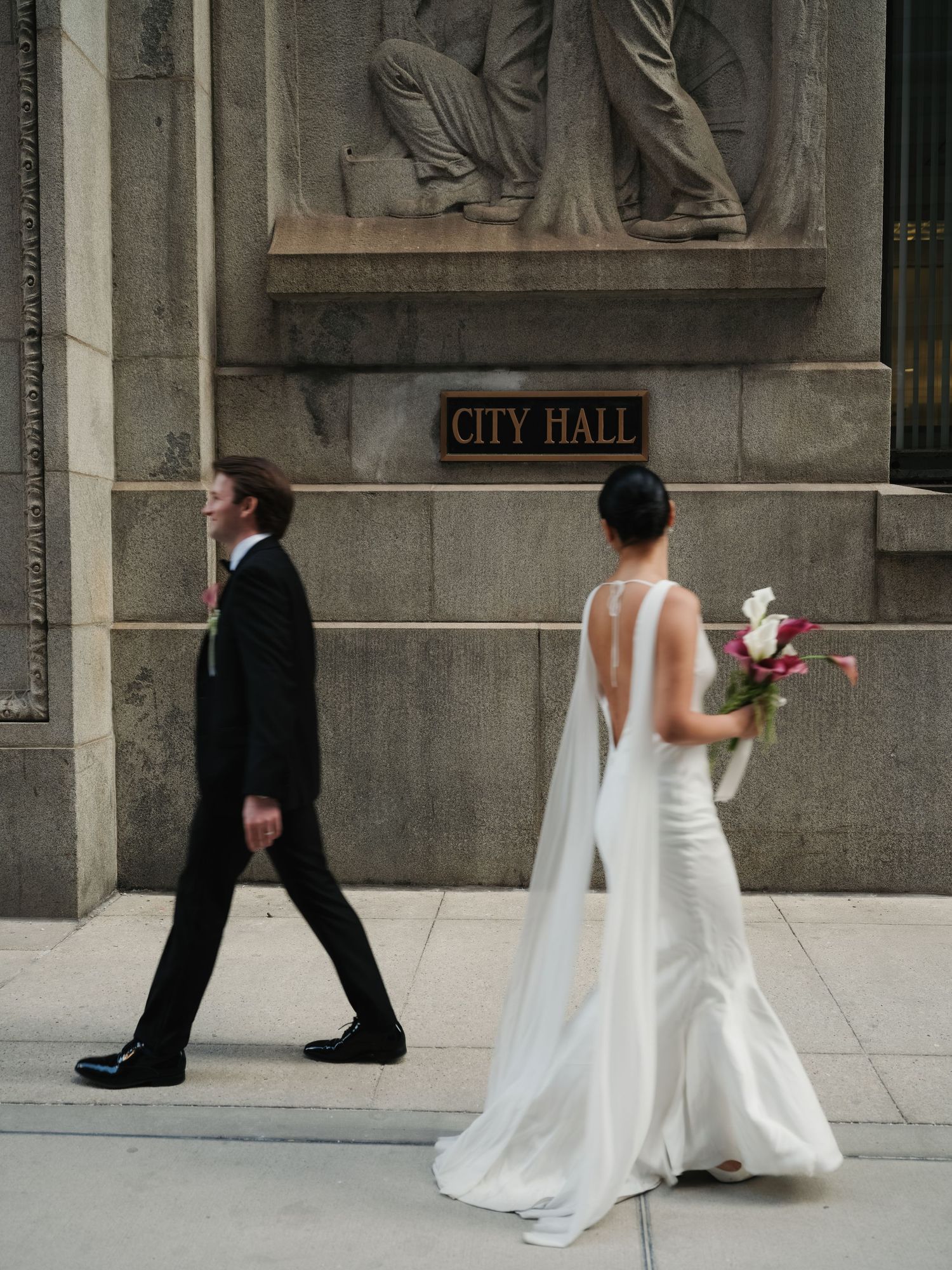 Bride and groom walking past City Hall exterior, bride in backless white gown carrying pink and white bouquet.