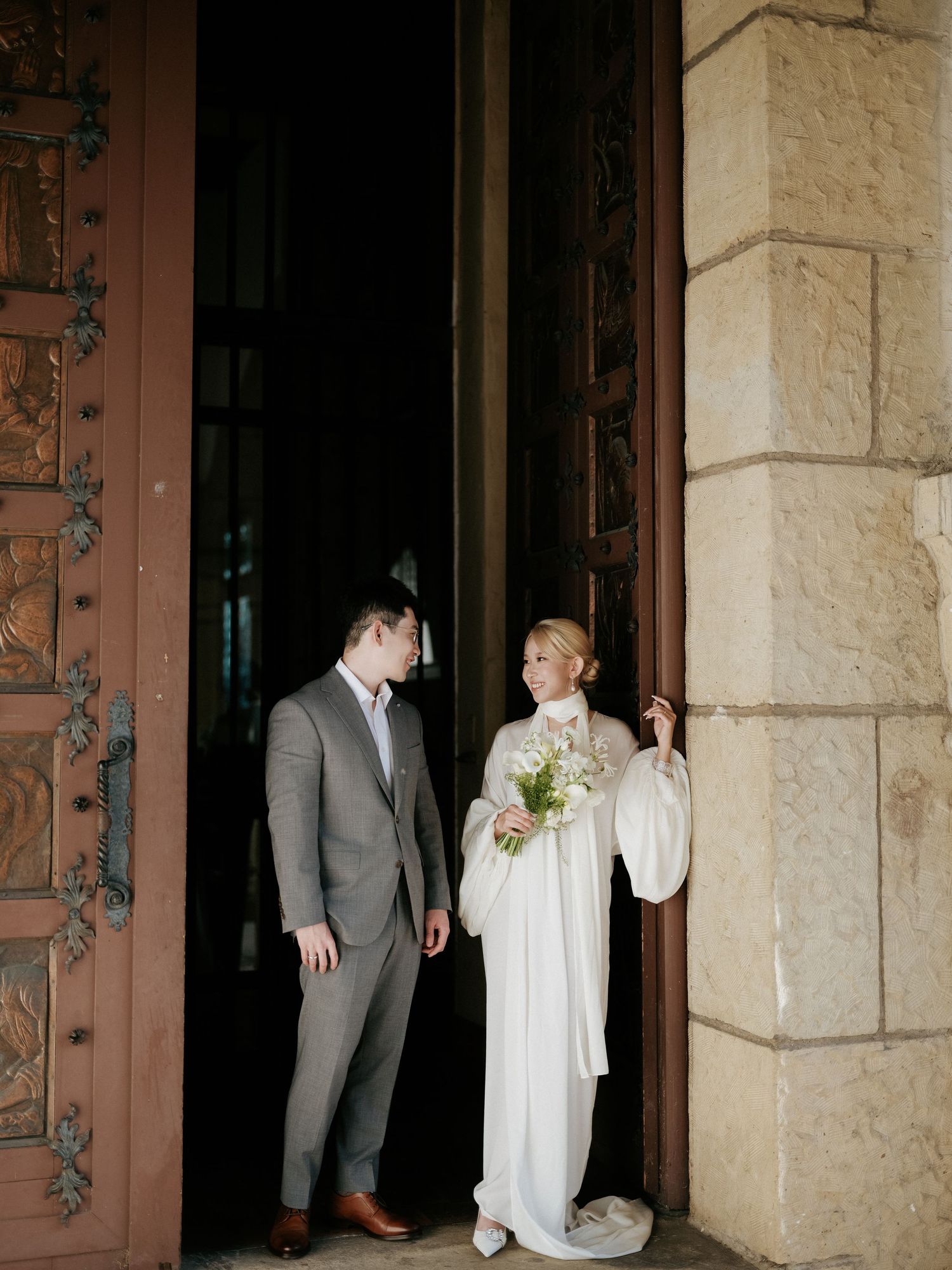Bride and groom share a glance beside ornate wooden doors of historic venue.