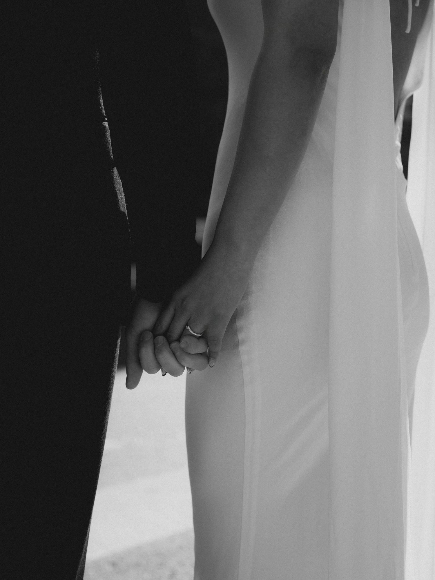 Black and white close-up of newlyweds holding hands, wedding ring visible, bride in white fitted gown.
