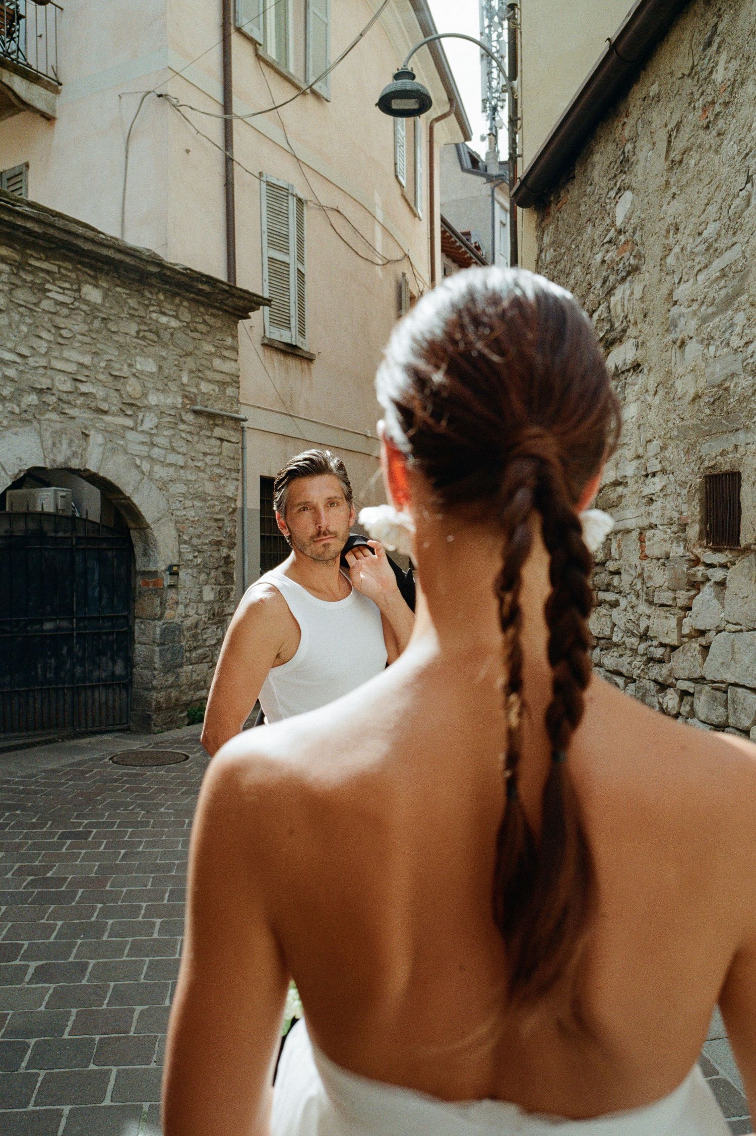 Groom in white tank top walks through a narrow European stone alley as bride with braided hair watches.