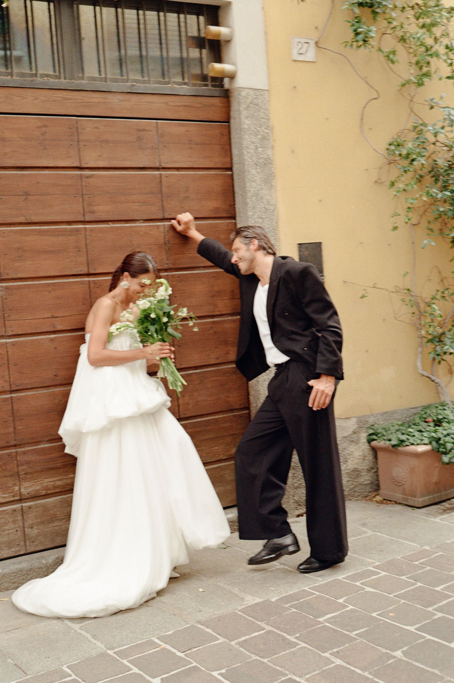 Groom in black suit leans over laughing bride in white gown outside rustic Italian building.