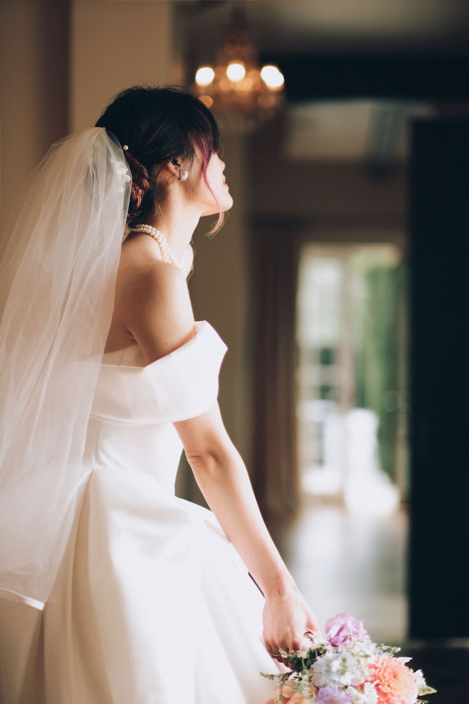 Bride in off-shoulder white gown with veil gazes upward holding colorful bouquet indoors.