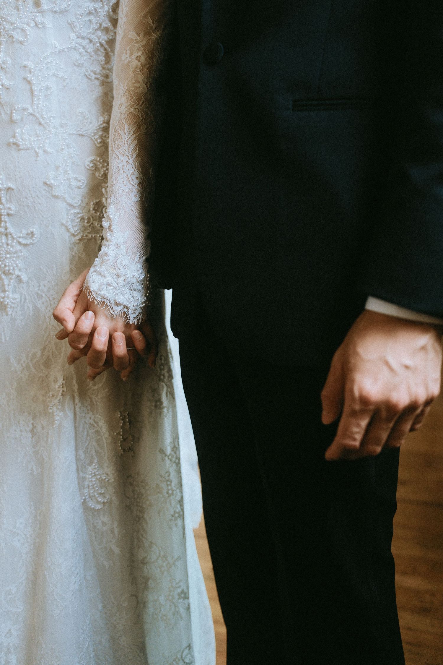 Close-up of bride in lace wedding gown and groom in black suit standing side by side holding hands.