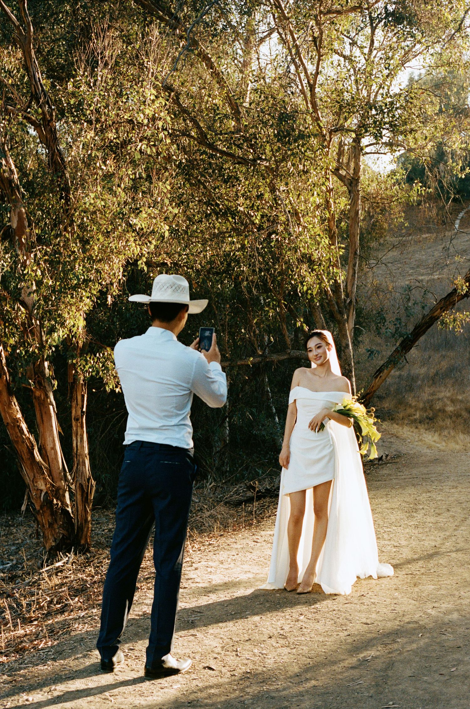 Groom in cowboy hat photographs smiling bride in off-shoulder white dress holding yellow bouquet outdoors.