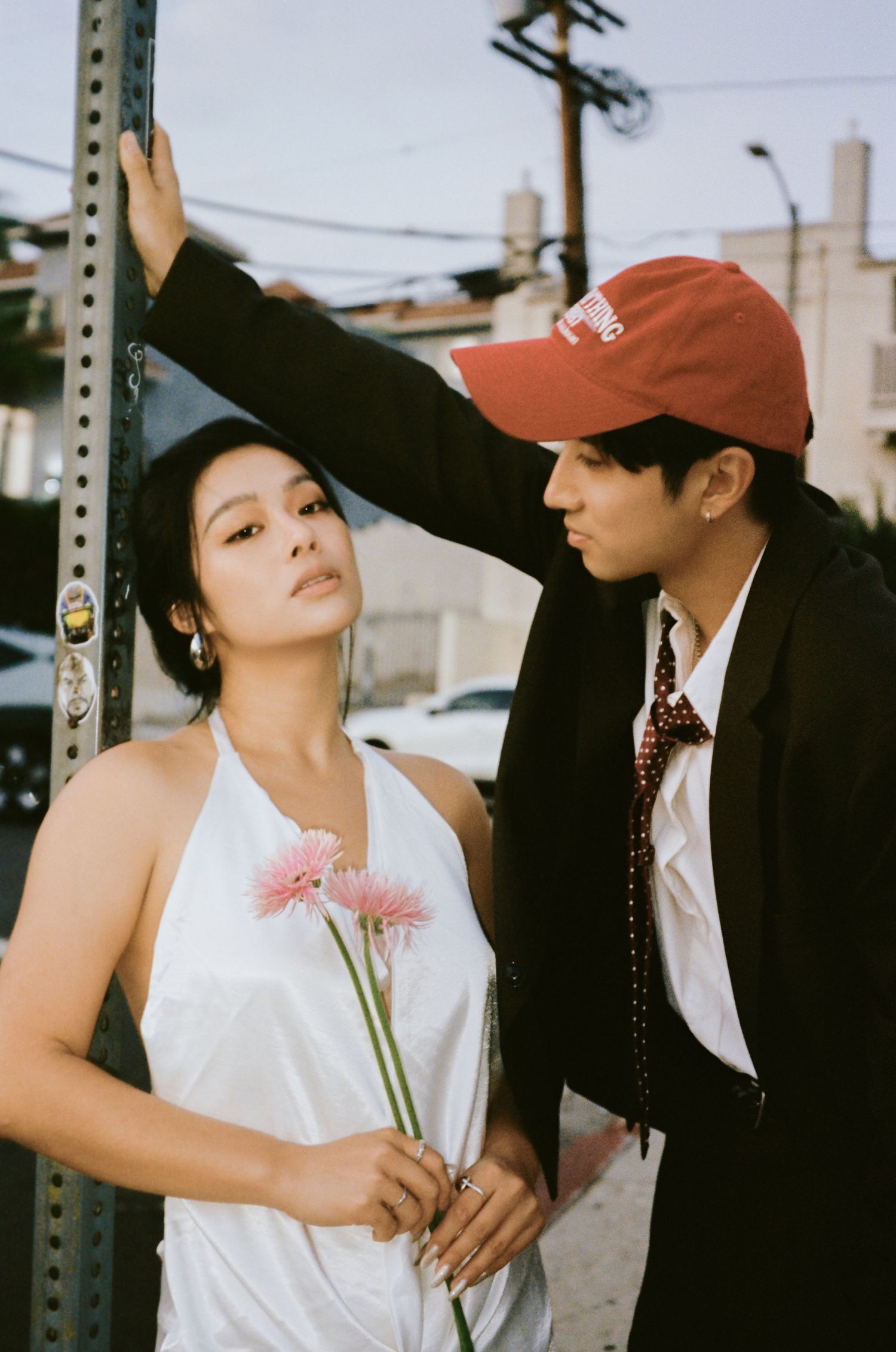 Couple posing on urban street, woman in white halter dress holding pink flowers, man in black blazer and red cap.