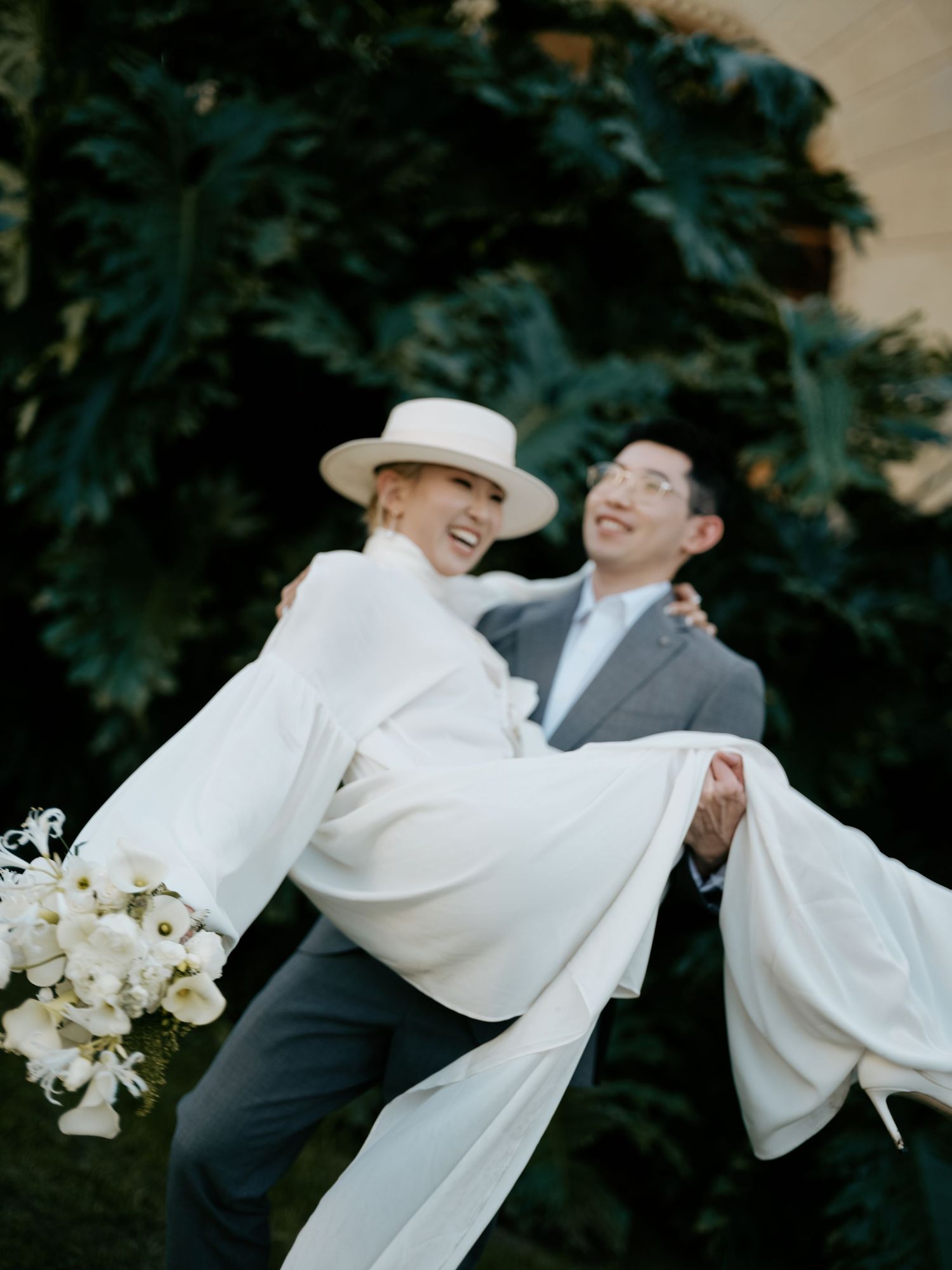 Joyful groom lifts bride in white flowing cape dress and boater hat outdoors.
