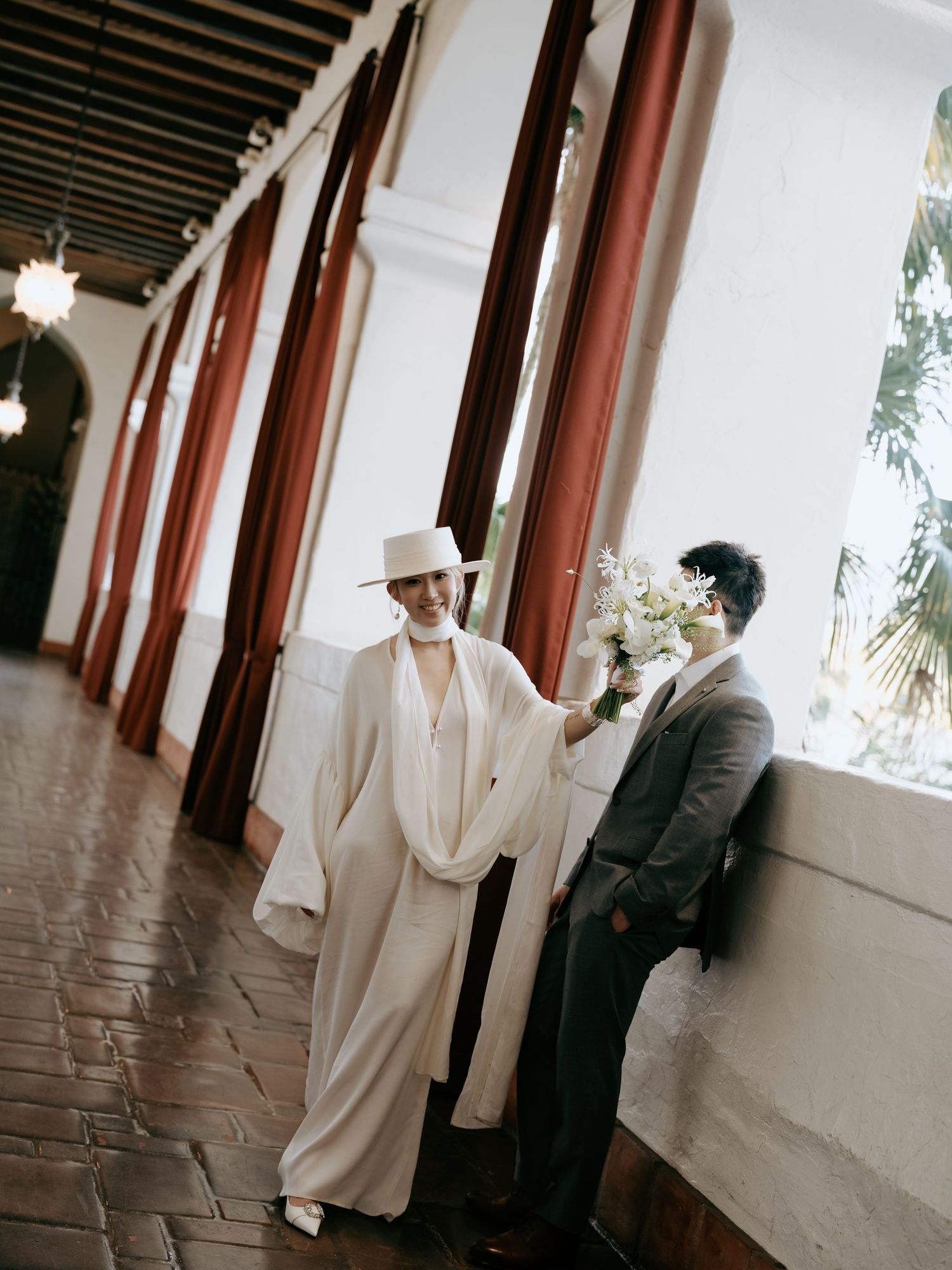 Bride in flowing white ensemble and hat smiles at groom leaning against wall in arcade.