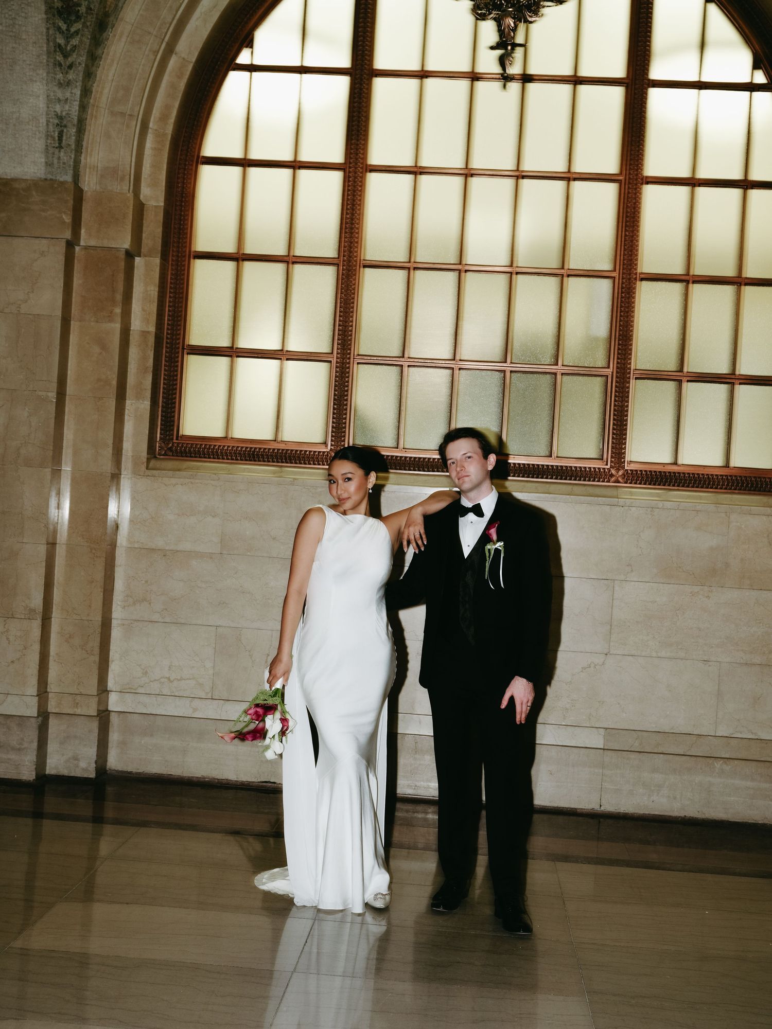 Bride and groom standing beneath arched window in elegant marble wedding venue interior.