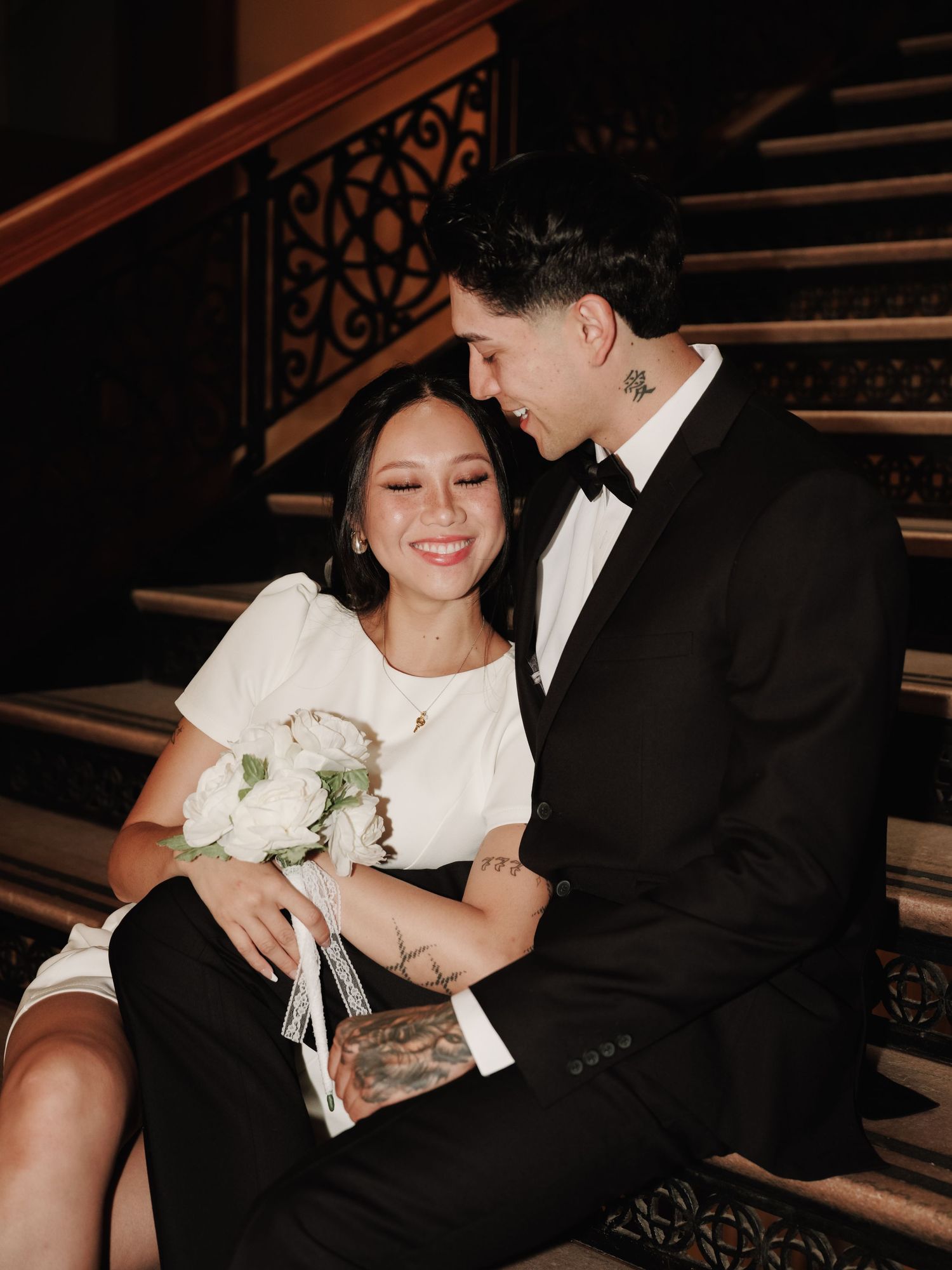 Happy bride holding white bouquet laughs with groom while sitting on grand staircase.