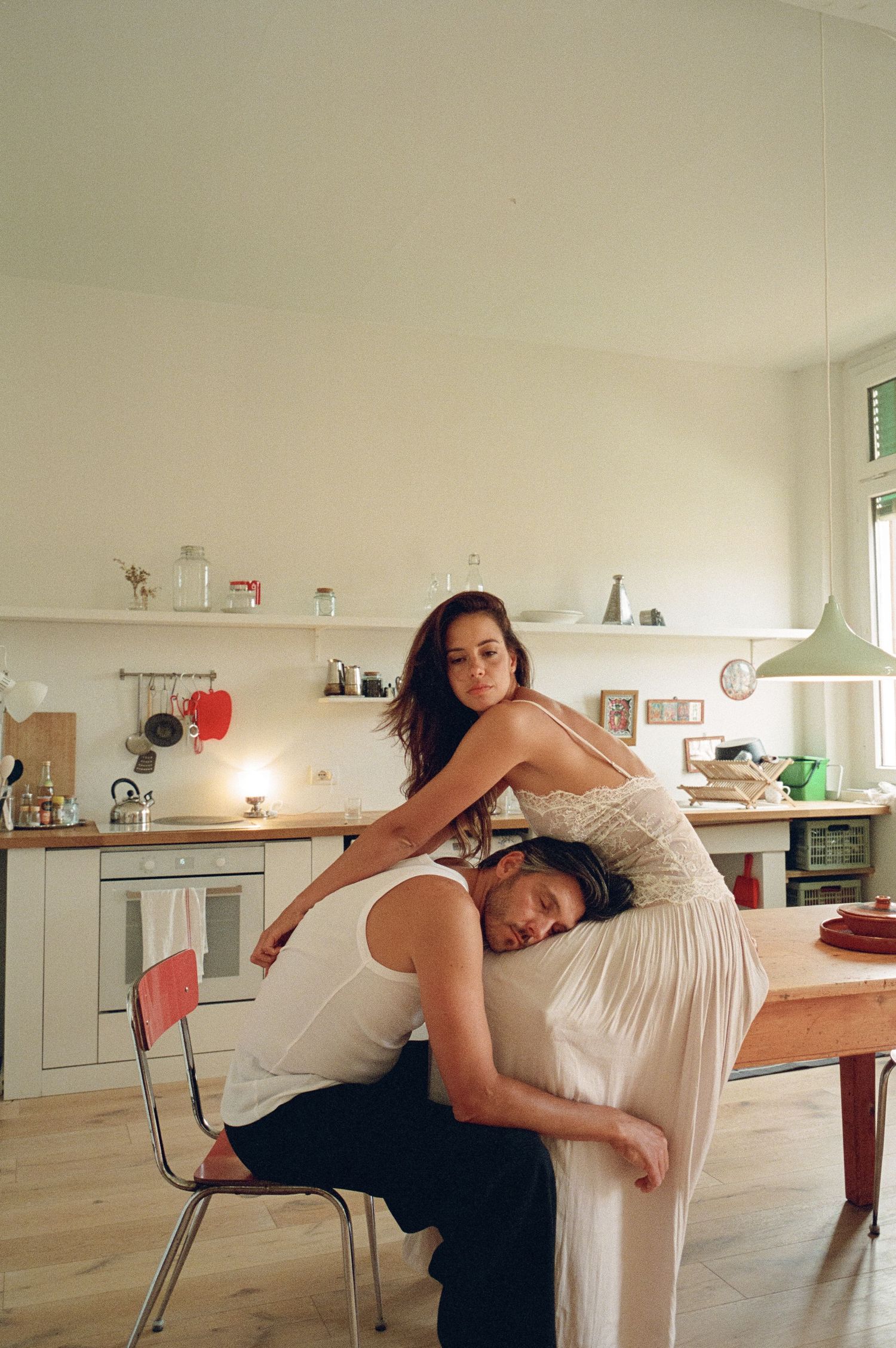 Bride in white slip dress resting on groom seated at a wooden kitchen table.