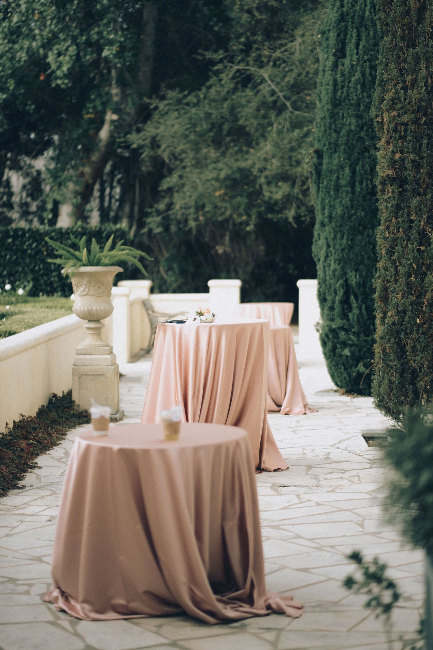 Outdoor wedding cocktail hour setup with blush pink draped round tables on a stone patio surrounded by cypress trees.