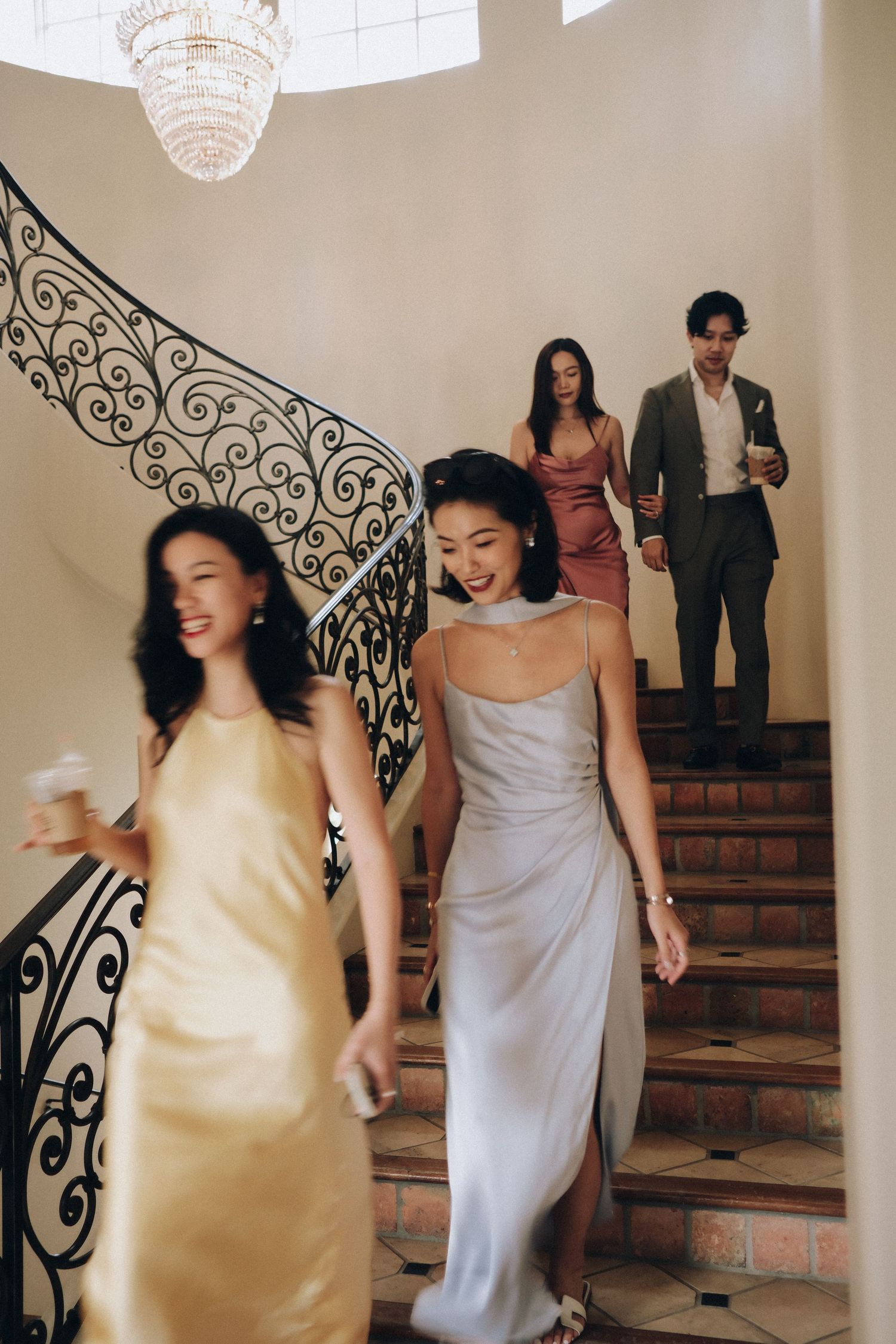 Elegantly dressed wedding guests descend ornate staircase with chandelier at reception venue.