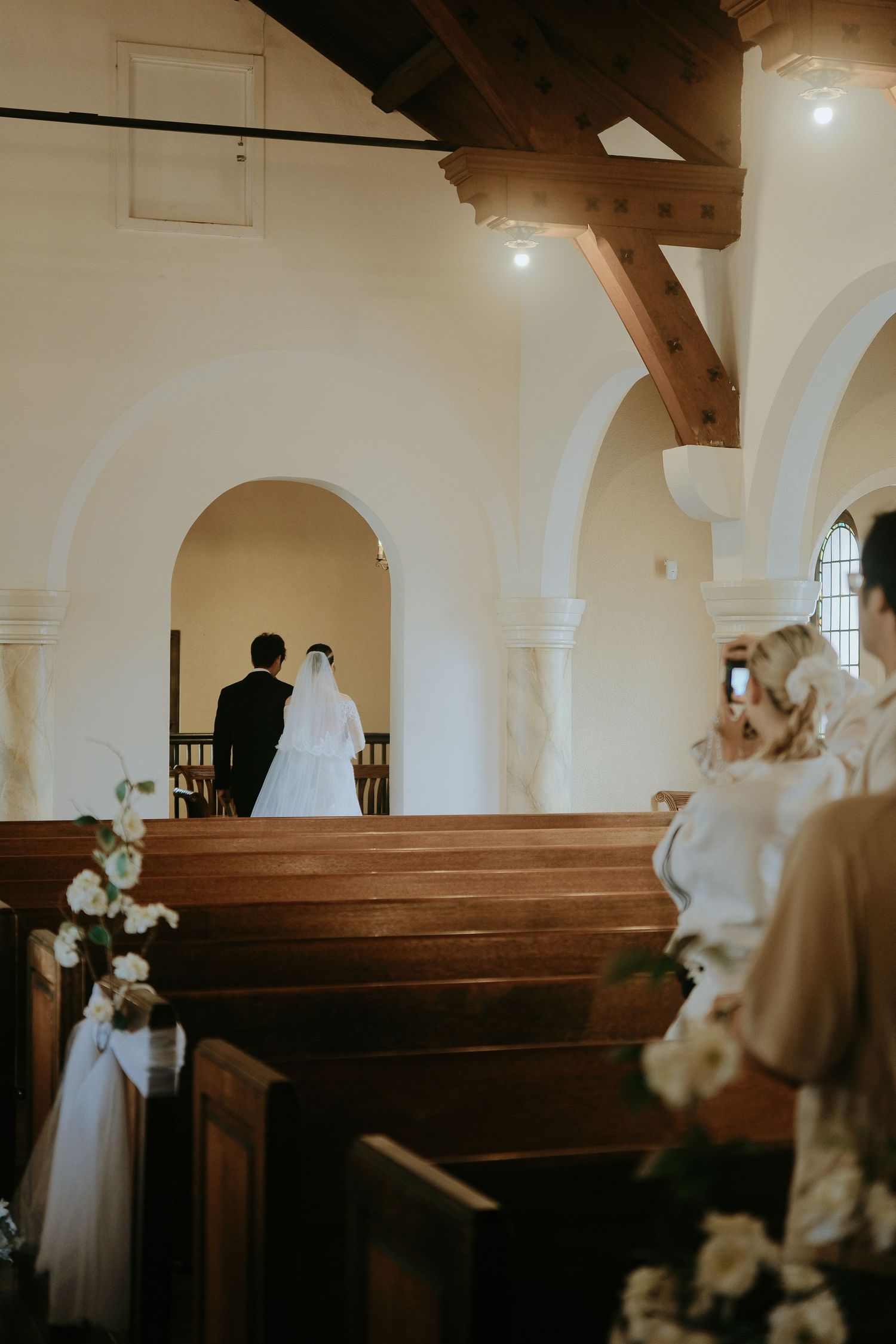 Bride and groom walk down the aisle of a white arched church with wooden pews and floral decorations.