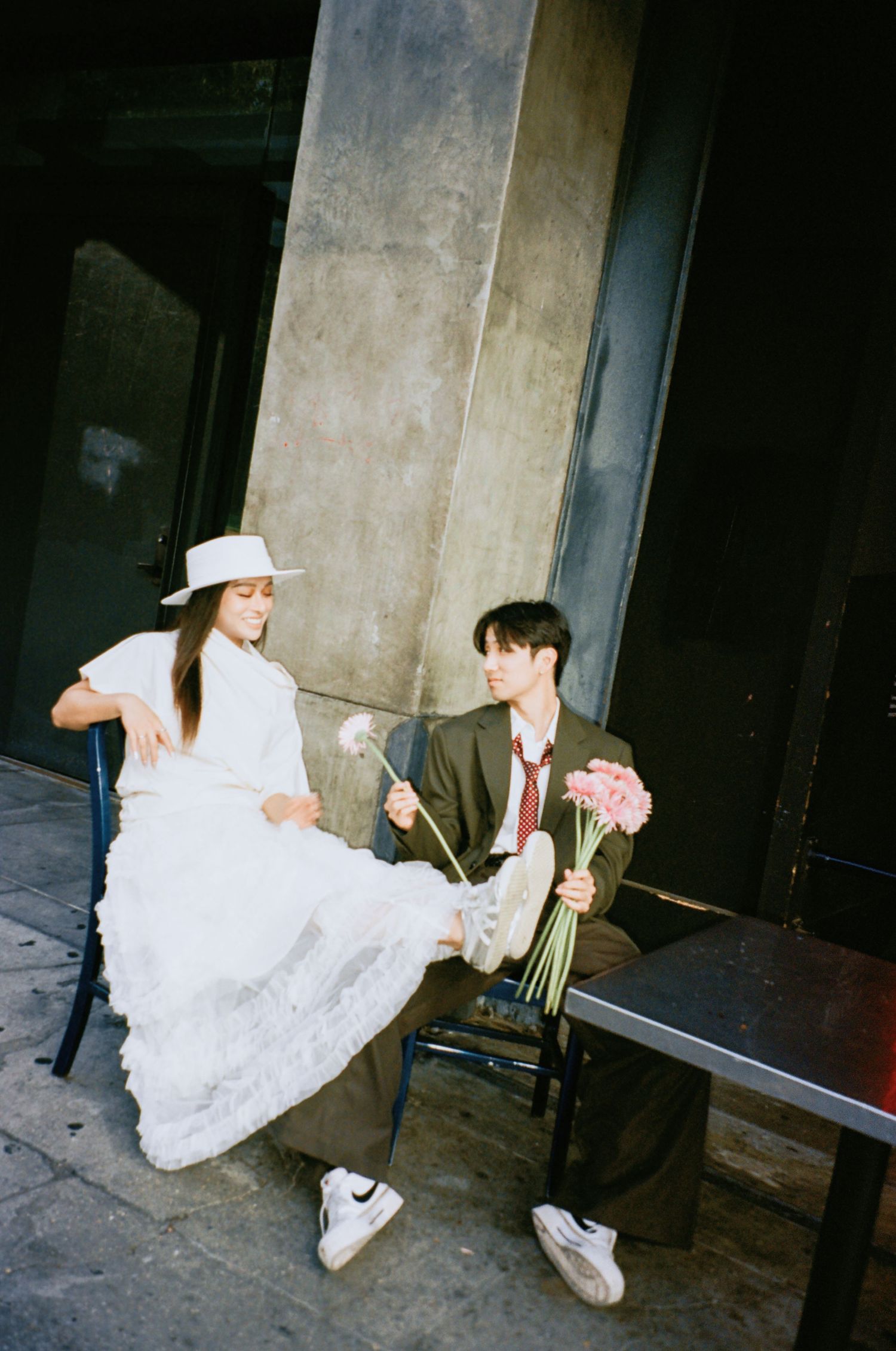 Couple sits at outdoor cafe table, woman in white lace dress and hat, man in olive suit holds pink flowers.