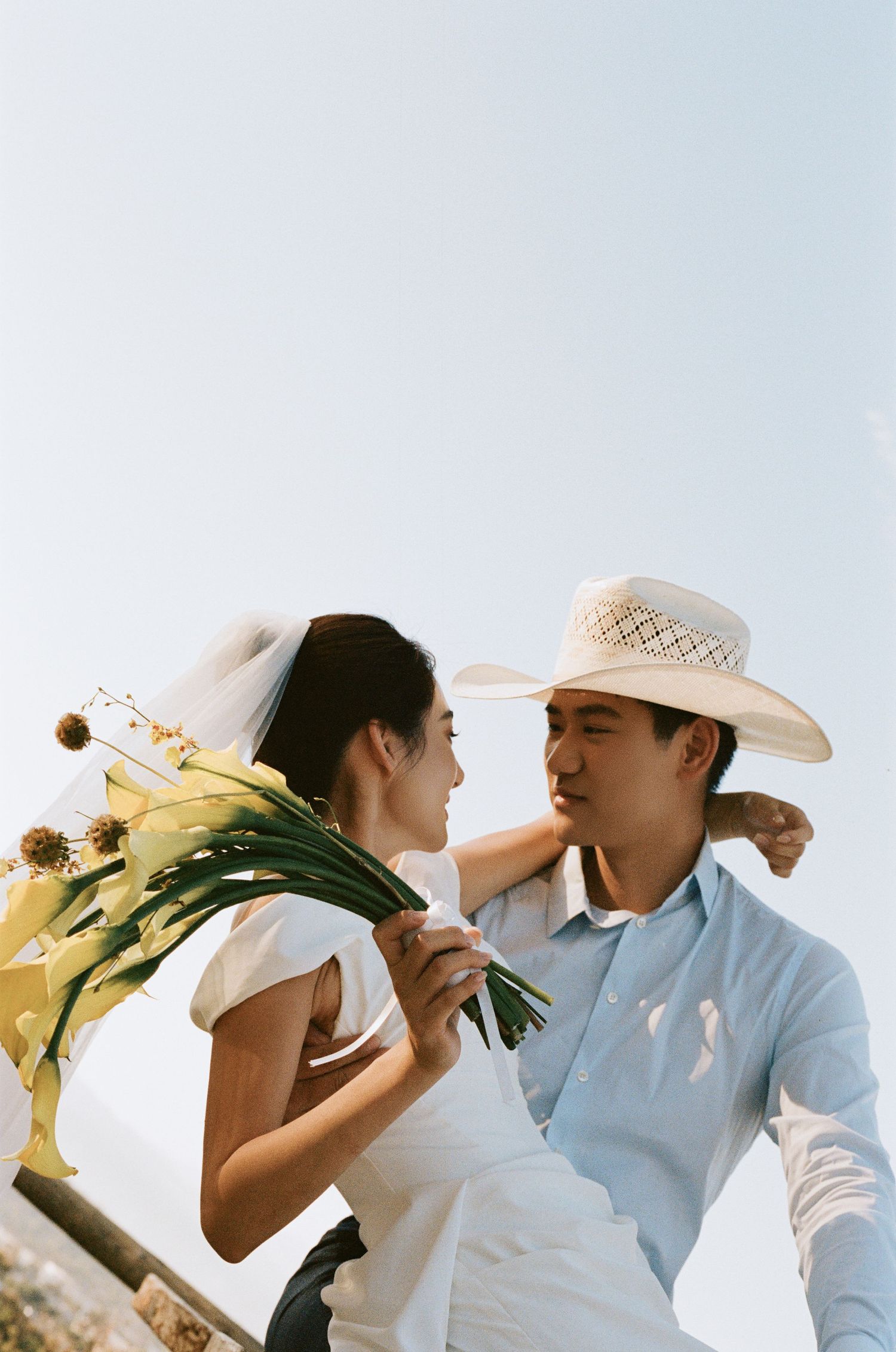 Smiling bride holding yellow flowers leans close to groom in cowboy hat under a clear sky.