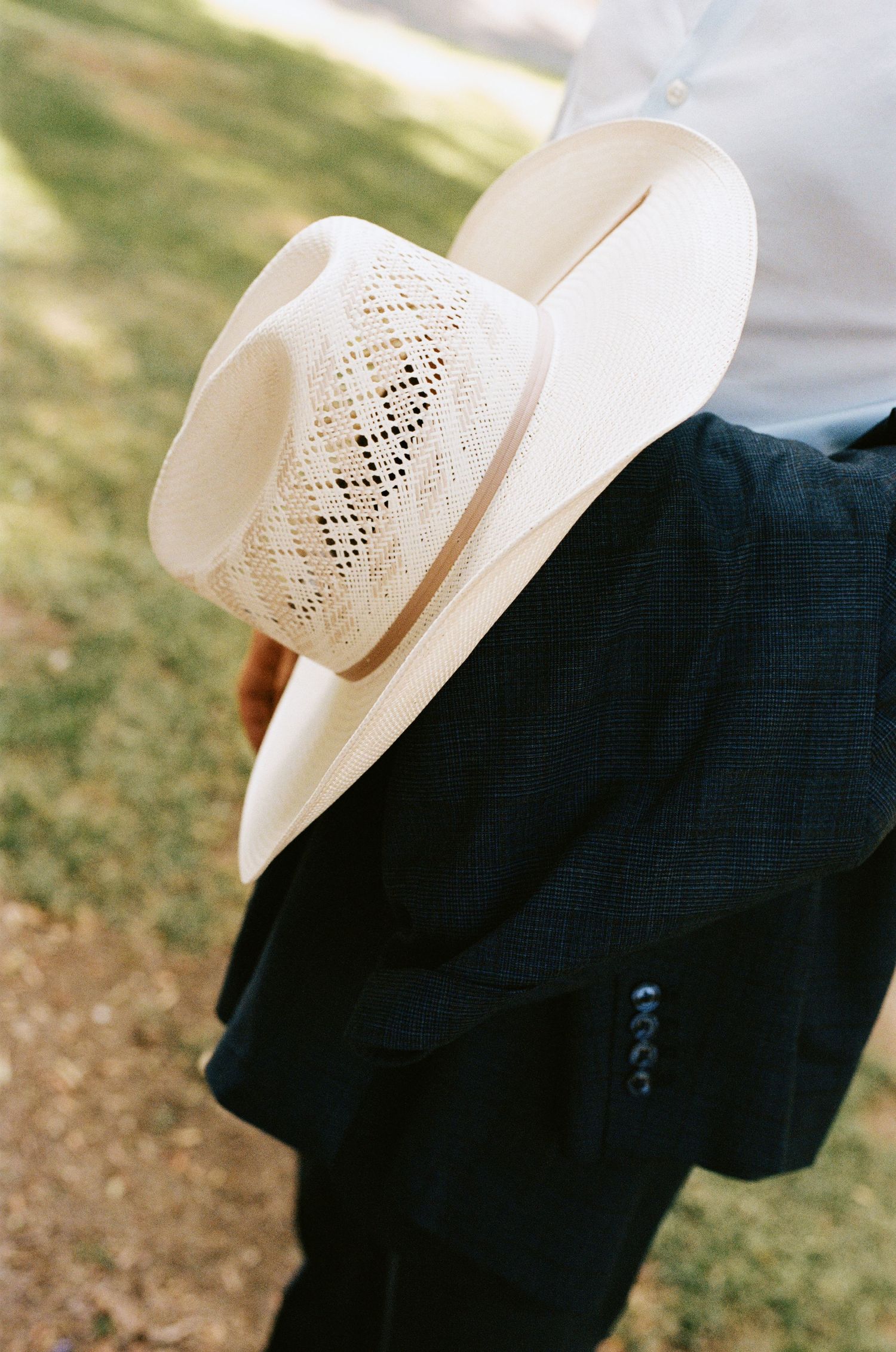 Groom in white shirt and dark trousers holds a cream woven cowboy hat against his side outdoors.