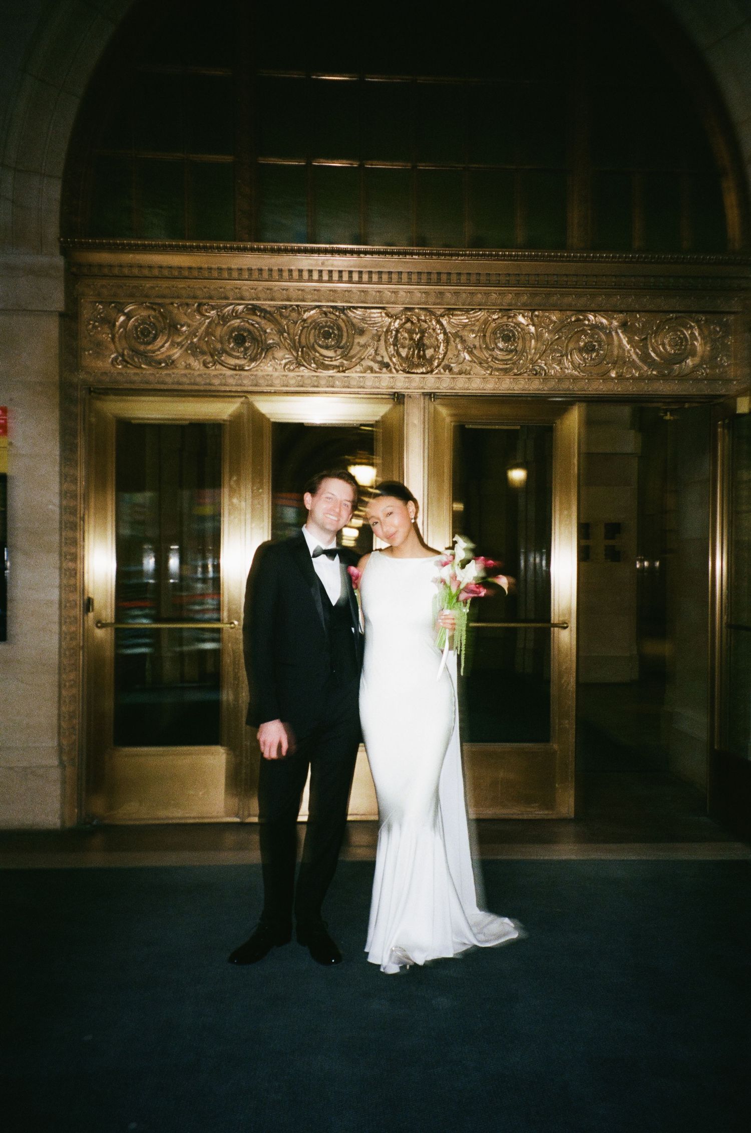 Bride and groom pose before grand gold ornate doors, she holds vibrant pink bouquet, he wears black tuxedo.