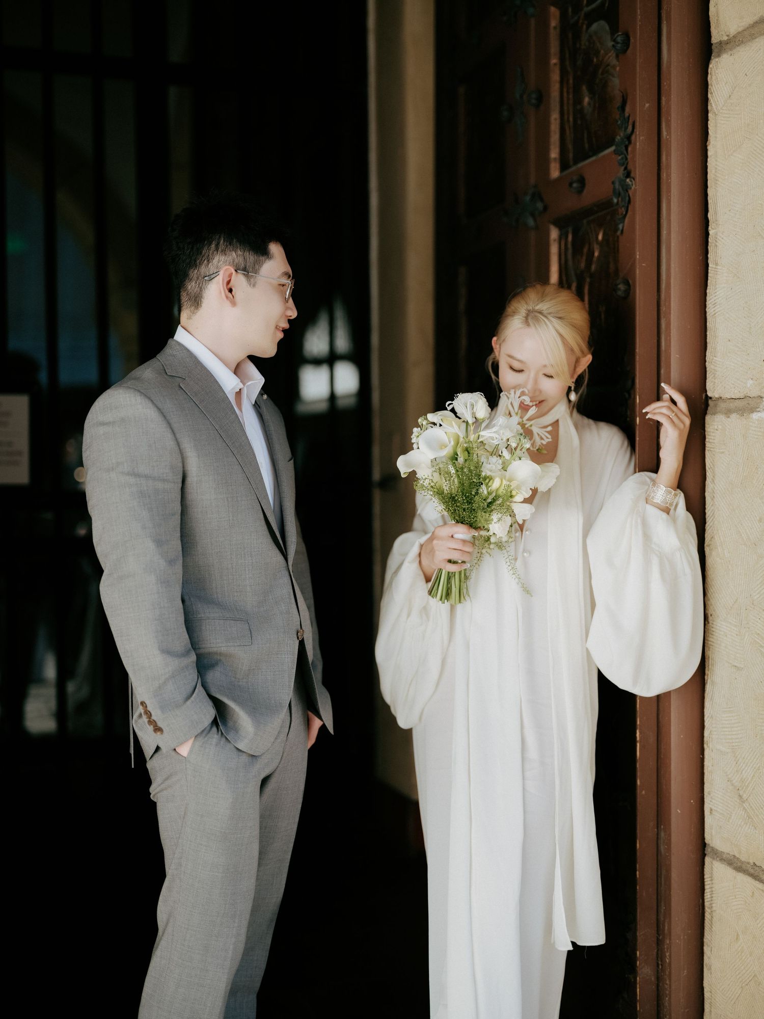 Bride in white gown with bouquet laughs with groom at historic building entrance.