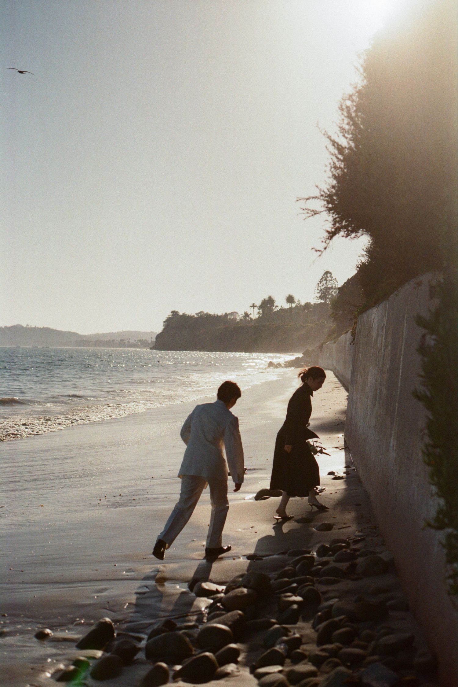 Couple walking along a sunlit rocky beach shoreline with coastal cliffs and calm ocean waves.