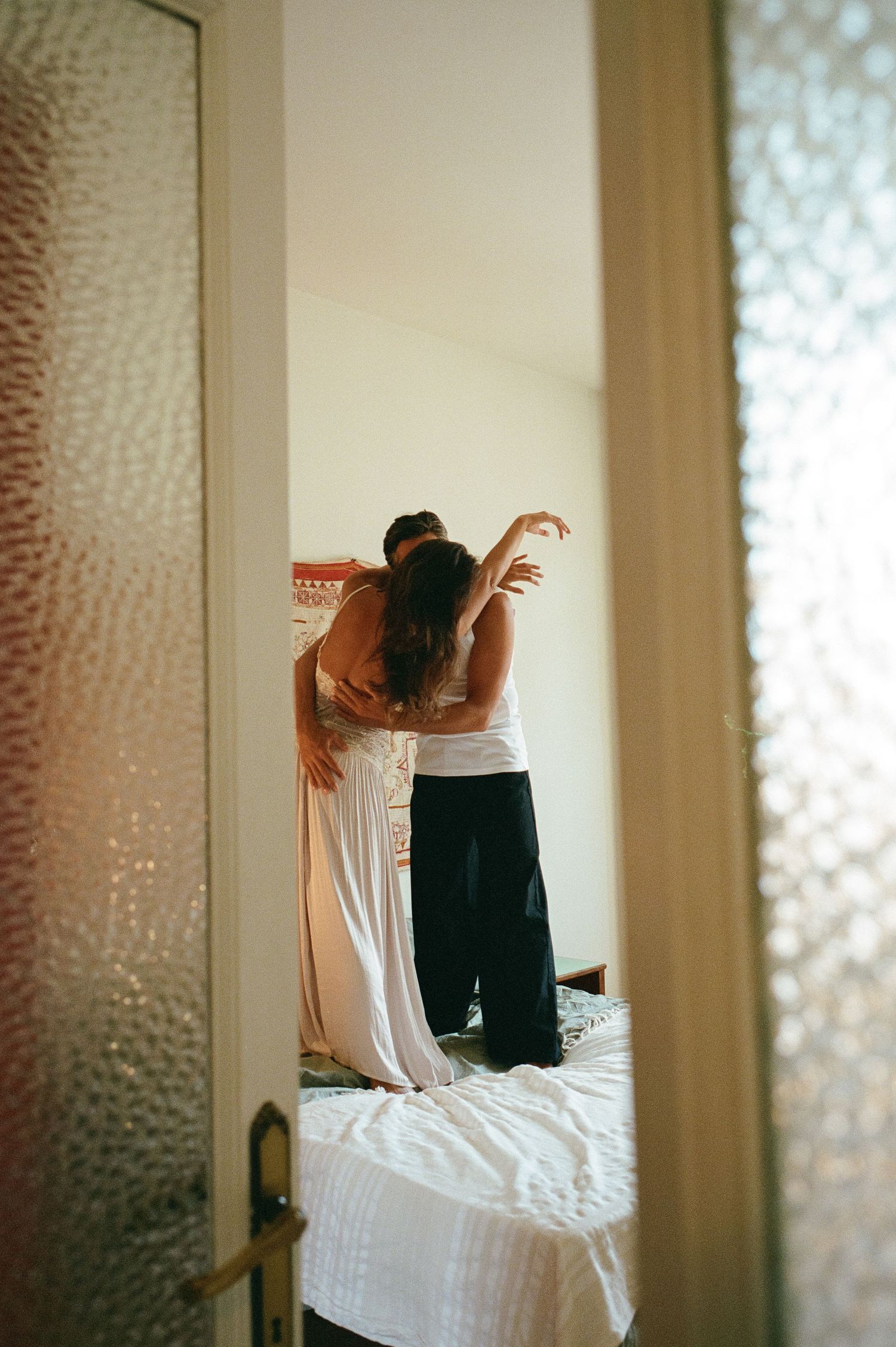 A couple embraces intimately on a bed, captured through a doorway with frosted glass panels in soft warm light.