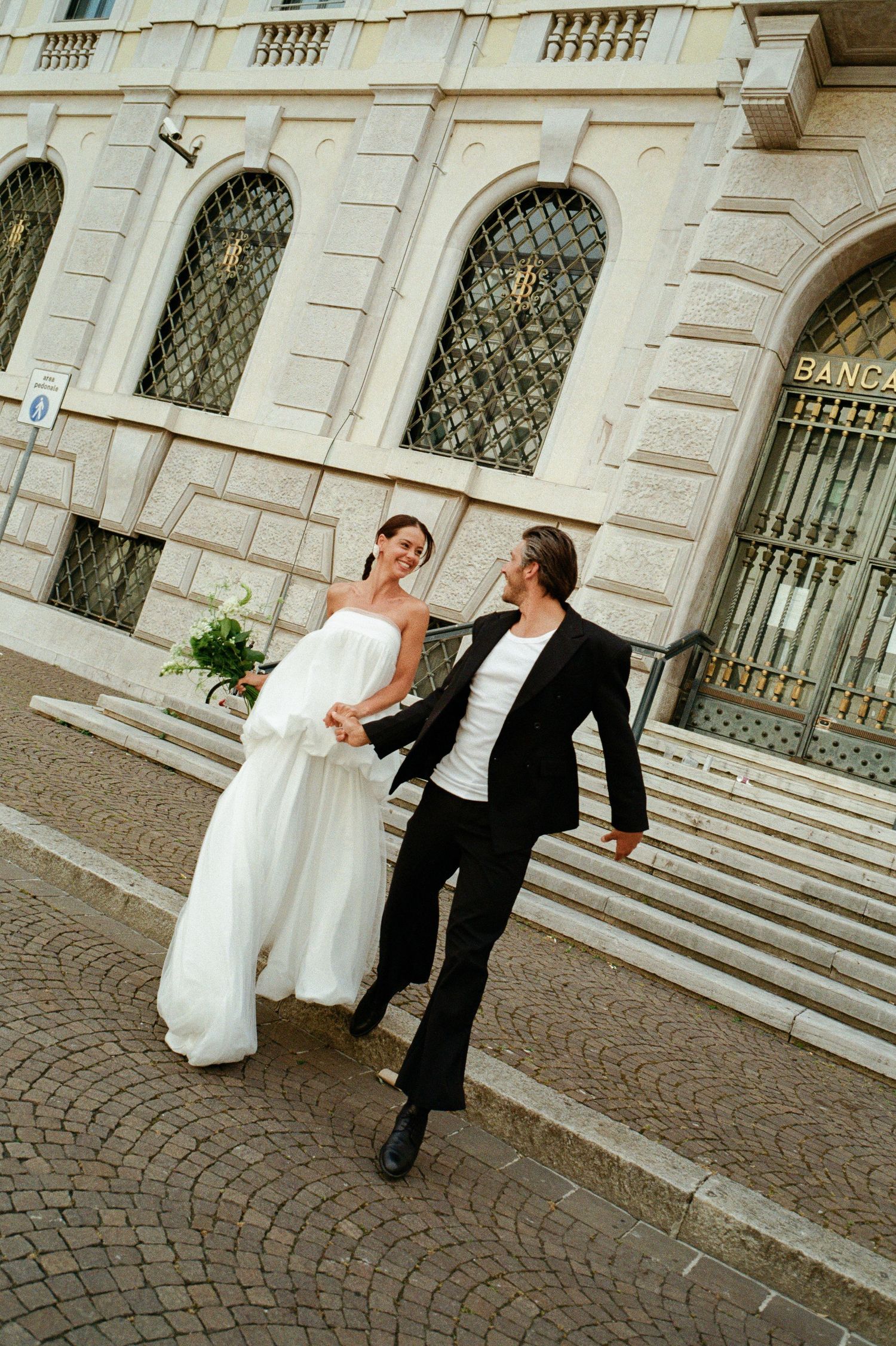 Bride in white gown and groom in black suit walk hand in hand laughing outside Italian bank building.