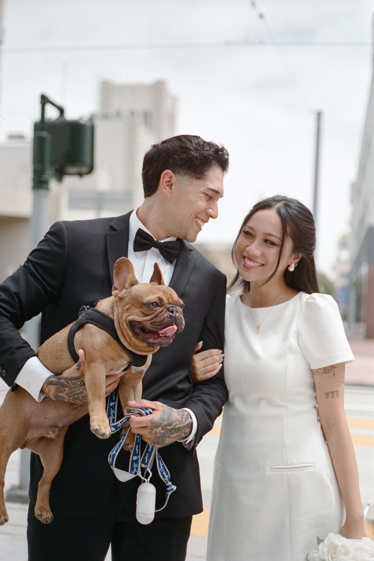 Groom in tuxedo holds French Bulldog while smiling at bride in white mini dress on city street.