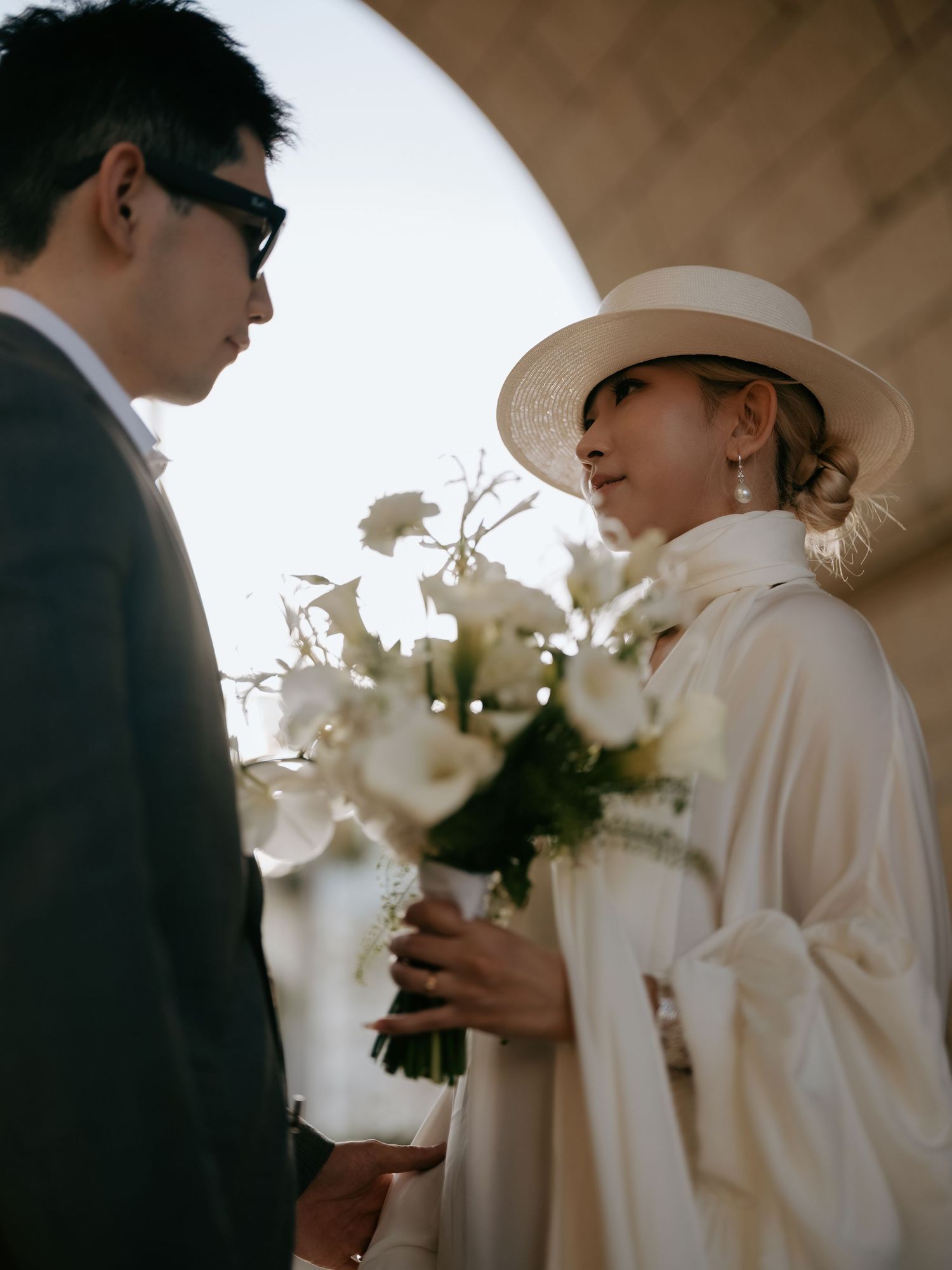 Bride in white boater hat holds white bouquet facing groom under stone archway.