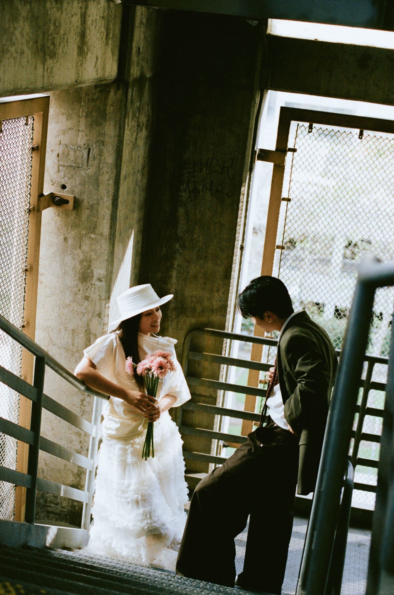 Bride in white ruffled dress and hat holds bouquet while groom looks down on an urban staircase.