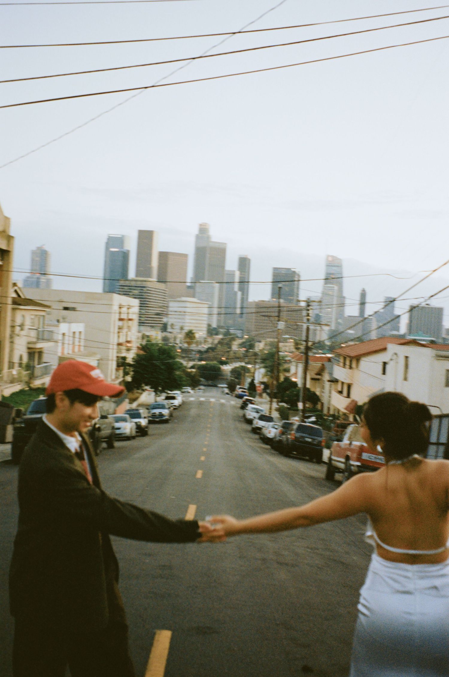 Couple holding hands on steep city street with downtown Los Angeles skyline in the background at dusk.