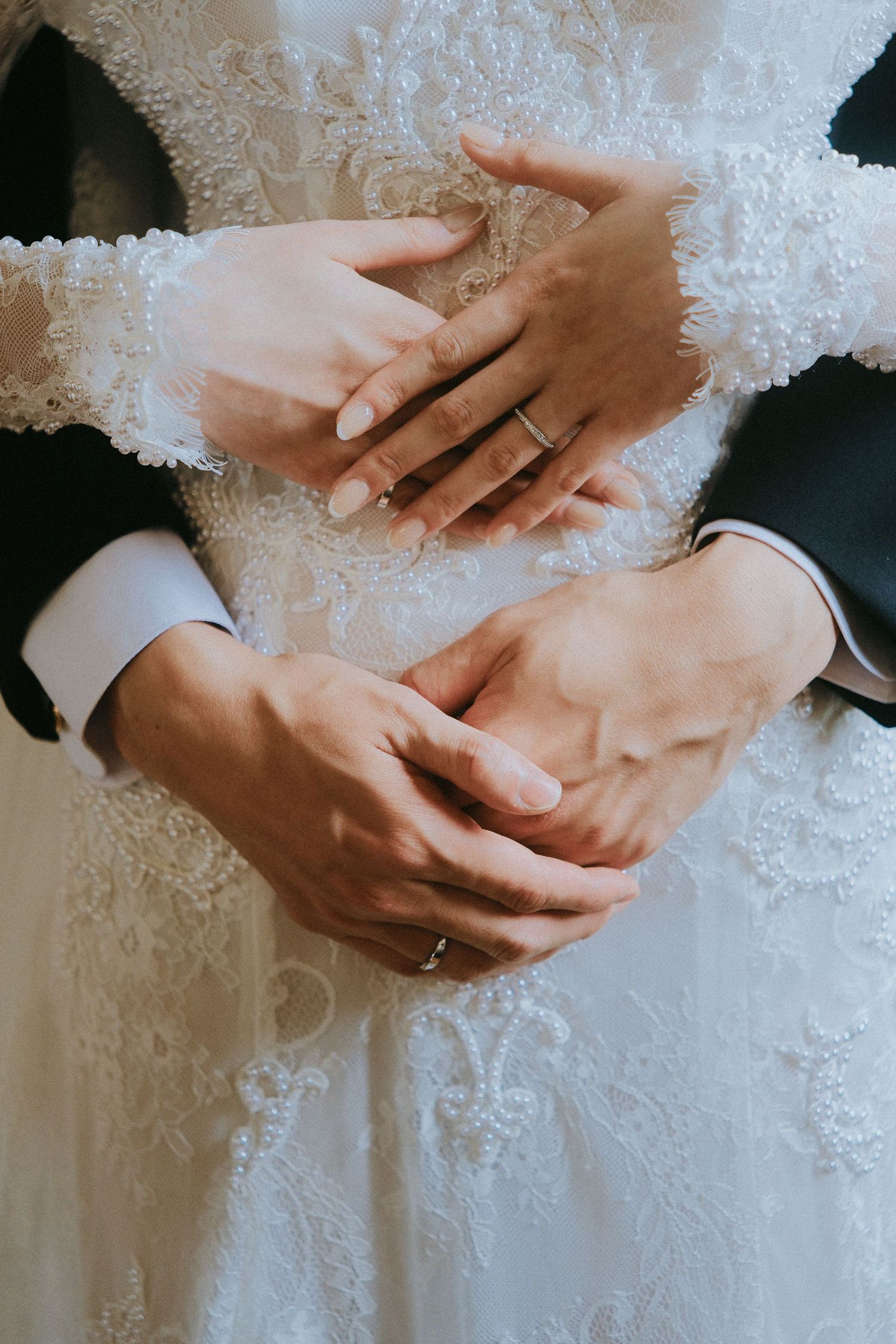 Close-up of bride and groom hands intertwined over intricate lace wedding dress with wedding rings.