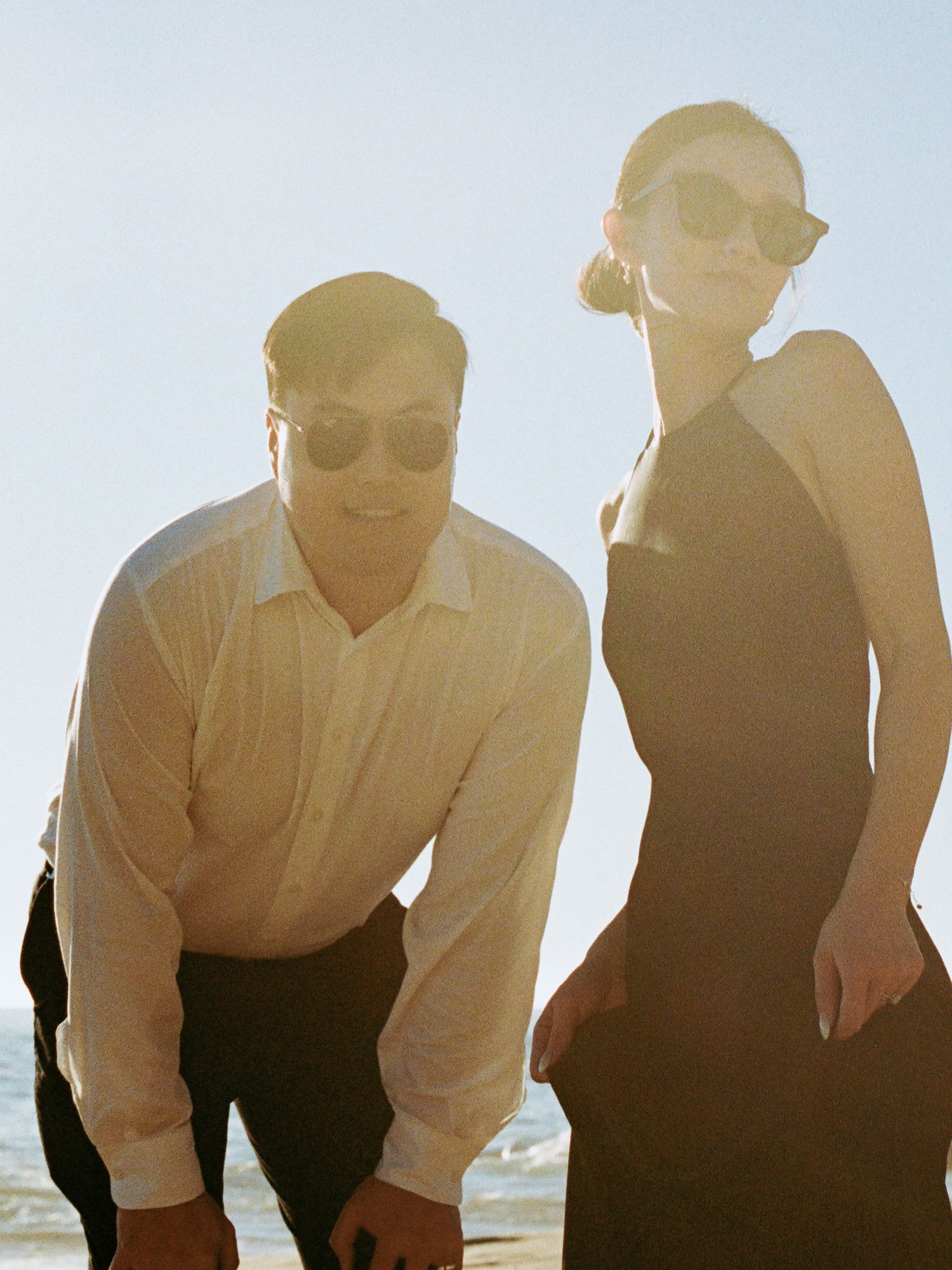 Couple wearing sunglasses smiling on sunny beach, man in white shirt and woman in black dress.