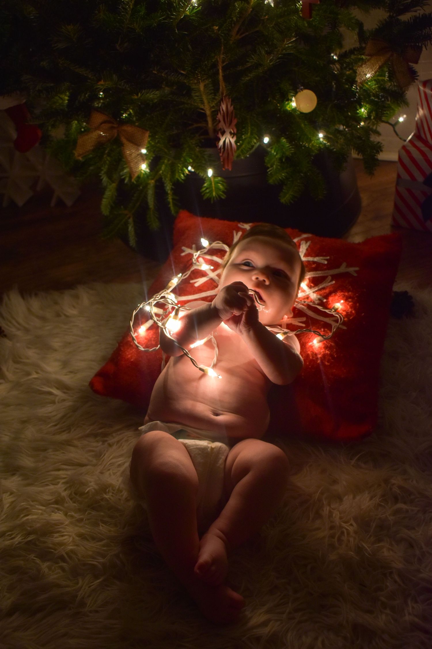 A baby lies under a Christmas tree wrapped in glowing string lights creating a magical holiday portrait.