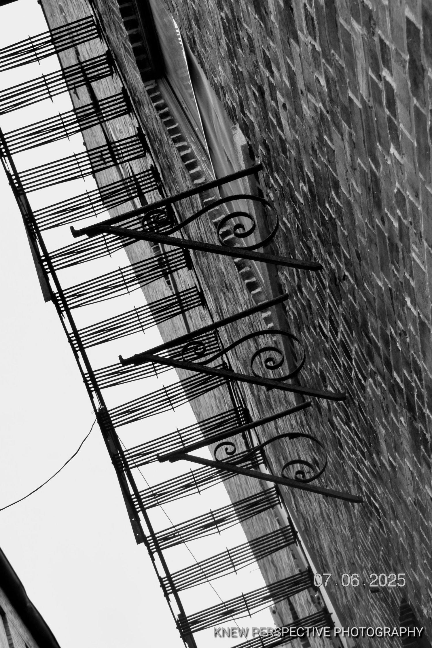 Black and white artistic photograph of stairs with ornate metal railings against a textured wall creating diagonal lines.