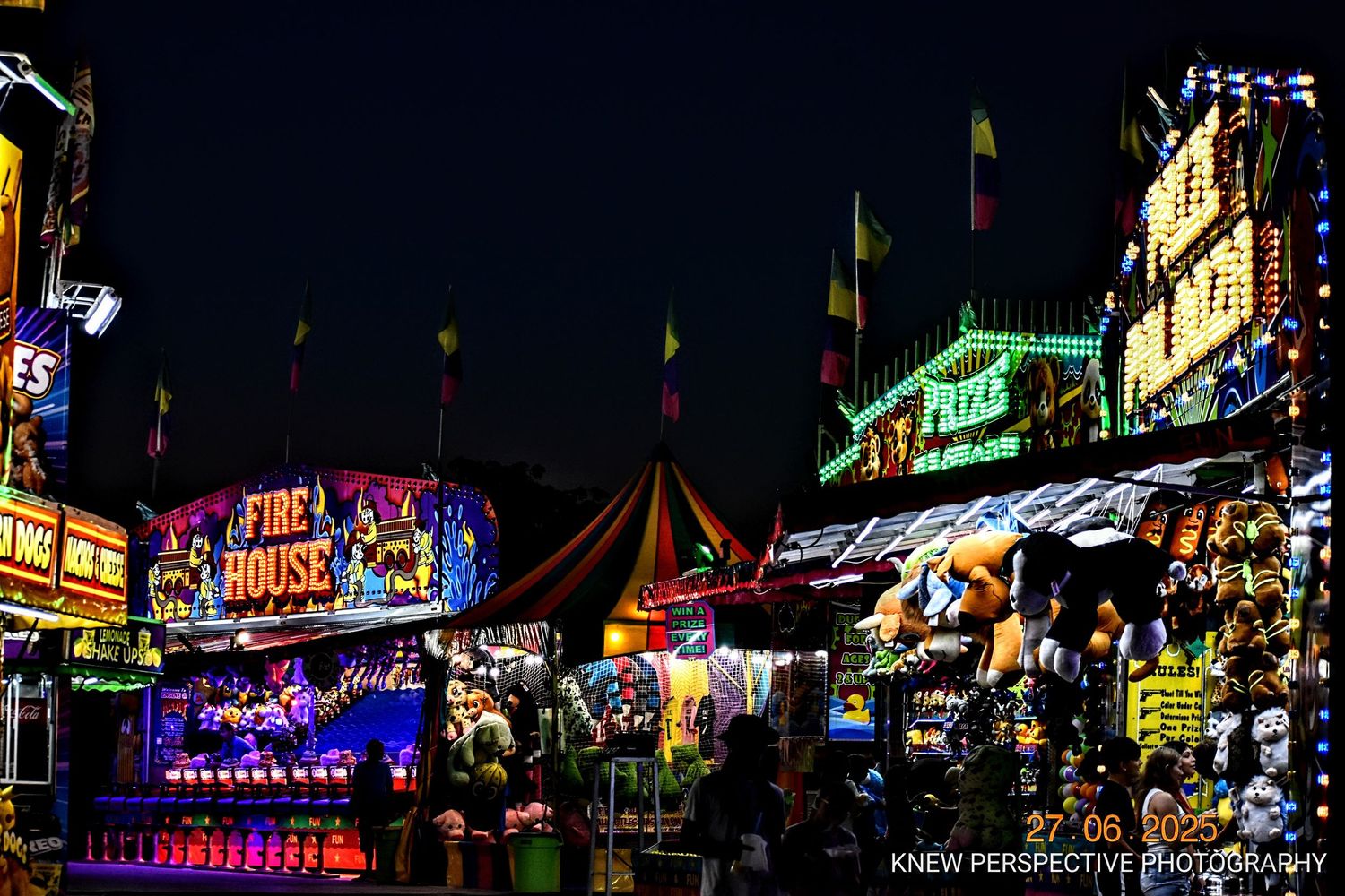 Colorful neon lights illuminate a bustling carnival midway with rides and food stands at night.