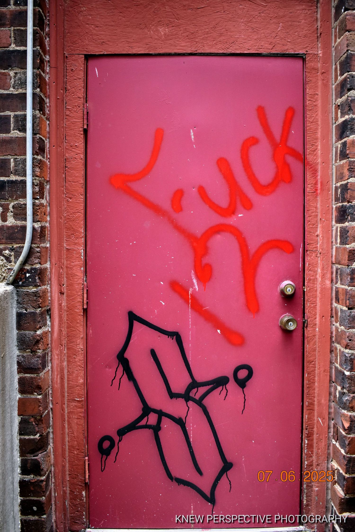 Red doors with black graffiti markings line up against a brick wall in an urban setting.