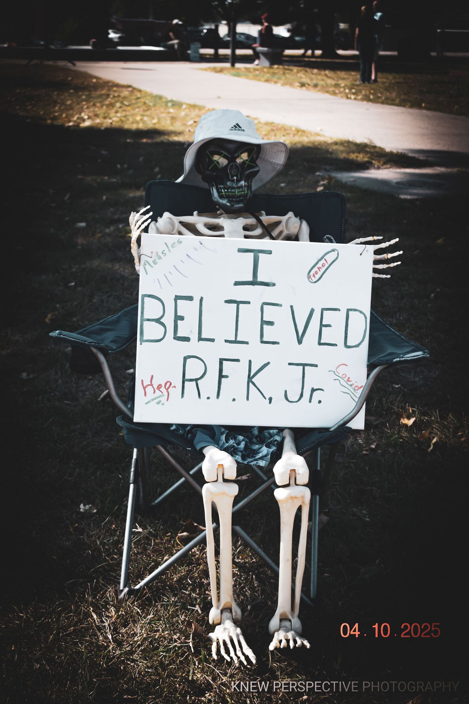 A skeleton decoration sits in a folding chair holding a sign that reads 'I BELIEVED R.F.K. Jr.' in a dark outdoor setting.