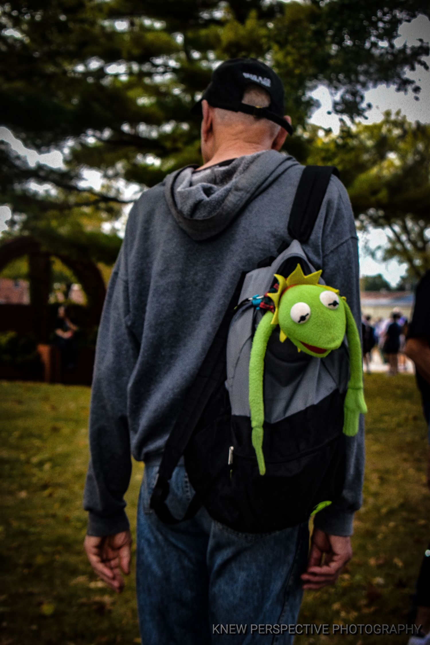 A small green Kermit the Frog plush toy peeks out from a dark backpack.