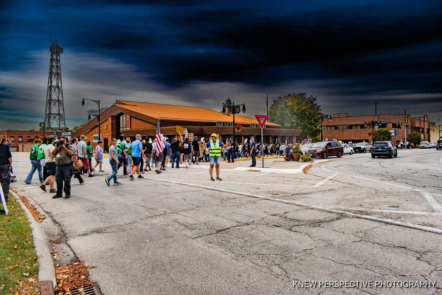 A long line of protesters marching across a crosswalk during the No Kings protest with a dark, cinematic stormy sky background.