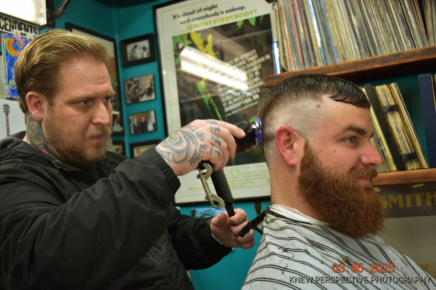 A barber with tattoos uses clippers to trim a client's hair in a shop with teal walls and vinyl records on display.