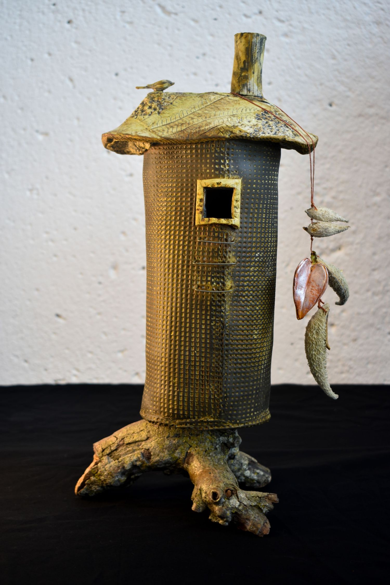 A rustic cylindrical house with textured metal surface and clay accents stands on branch-like feet against a white wall.