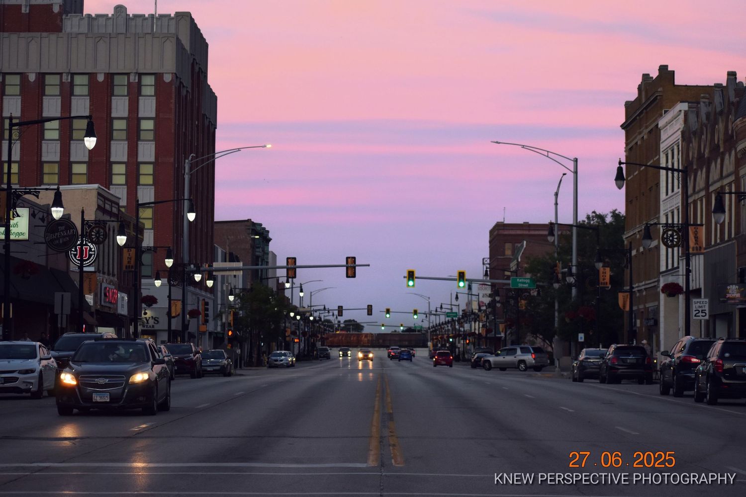A city street at sunset with brick buildings and glowing streetlights casting warm light against a pink and purple sky.