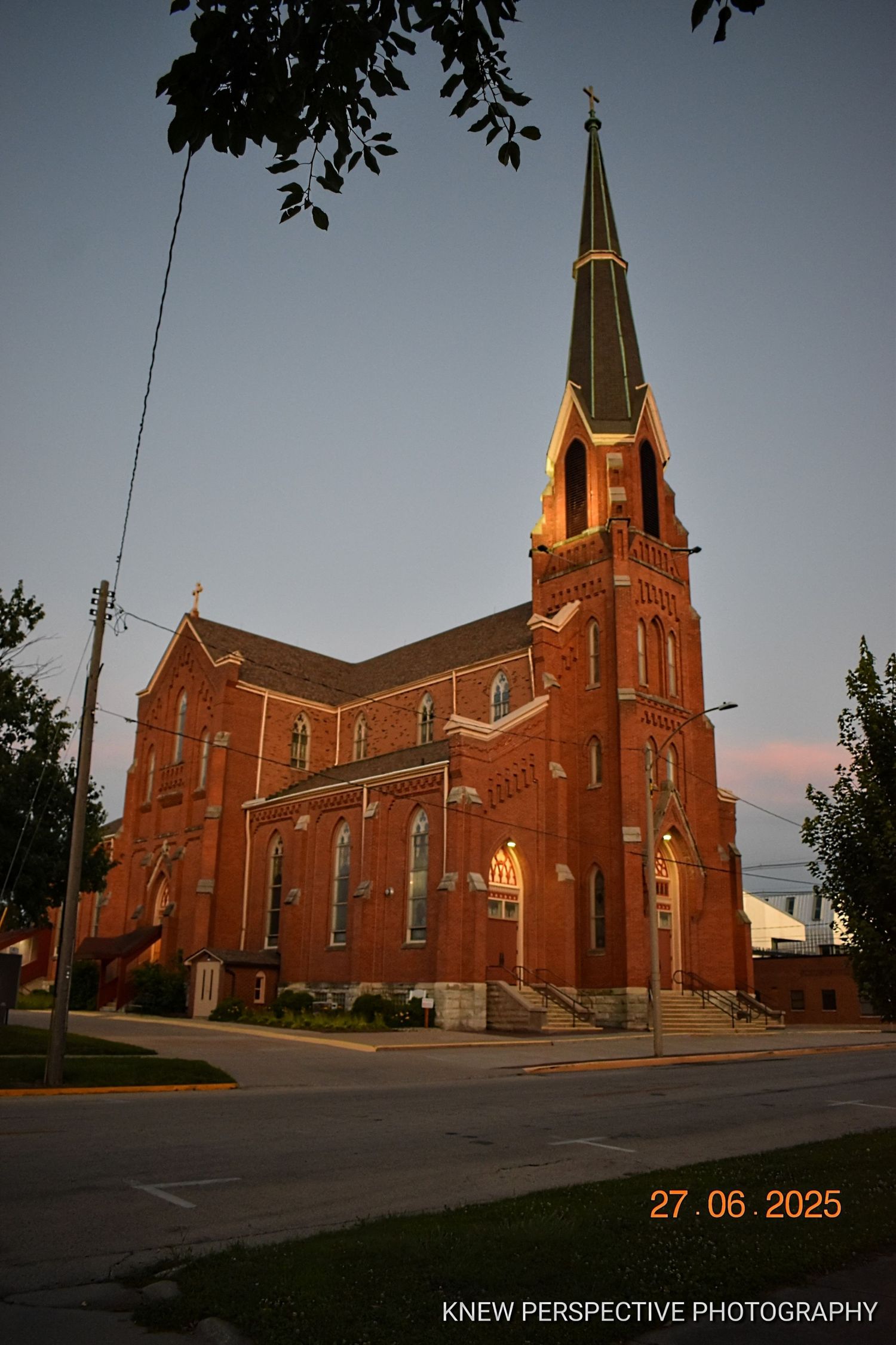 A red brick Gothic church with tall spire illuminated at dusk, photographed from a street corner perspective.