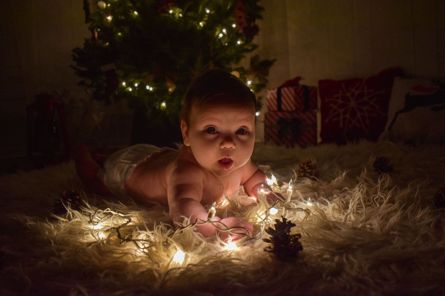 A baby lying on a white blanket surrounded by glowing Christmas lights with a decorated tree in the background.