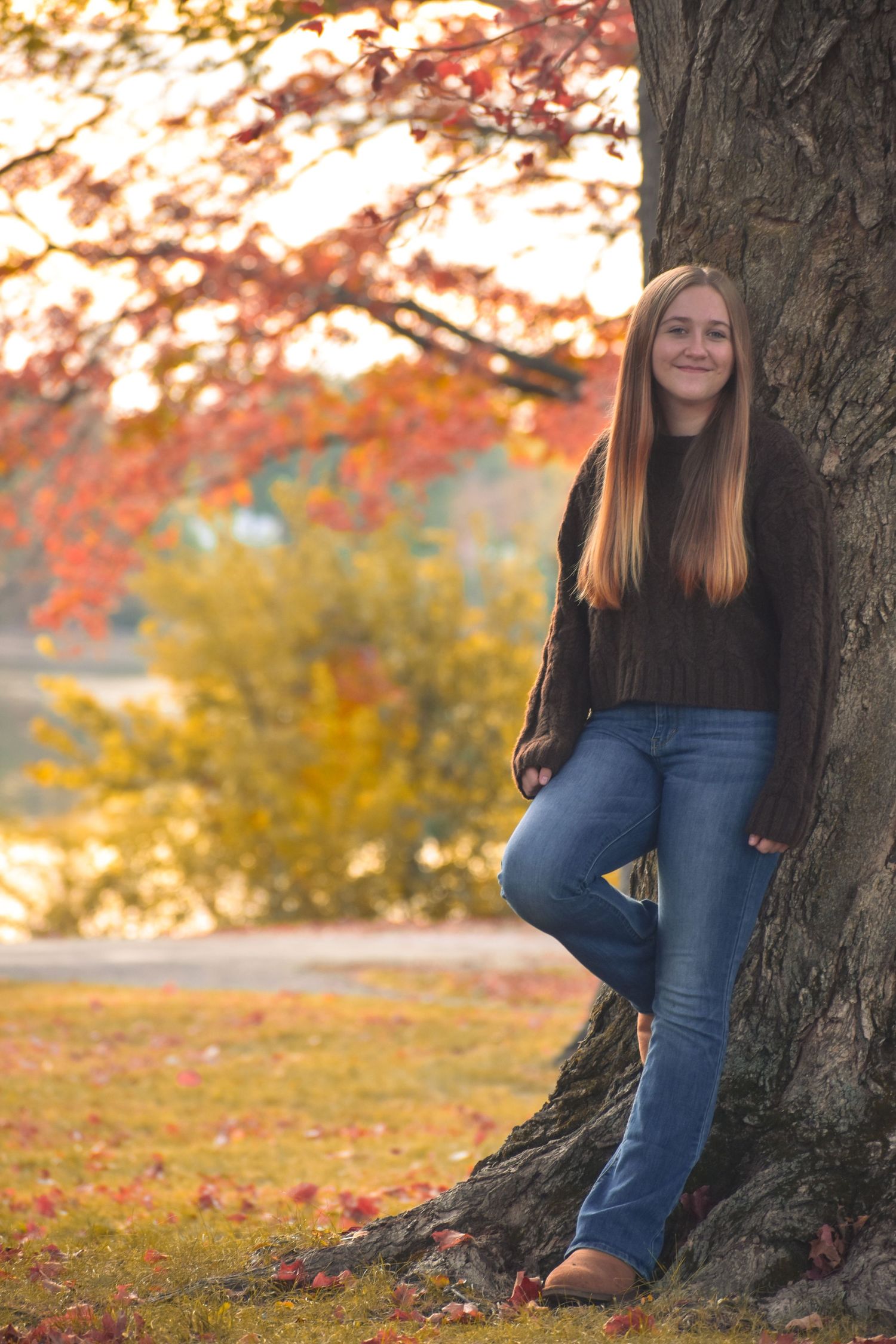 A person in jeans and brown boots leans against a large tree trunk during autumn with colorful fall foliage in the background.